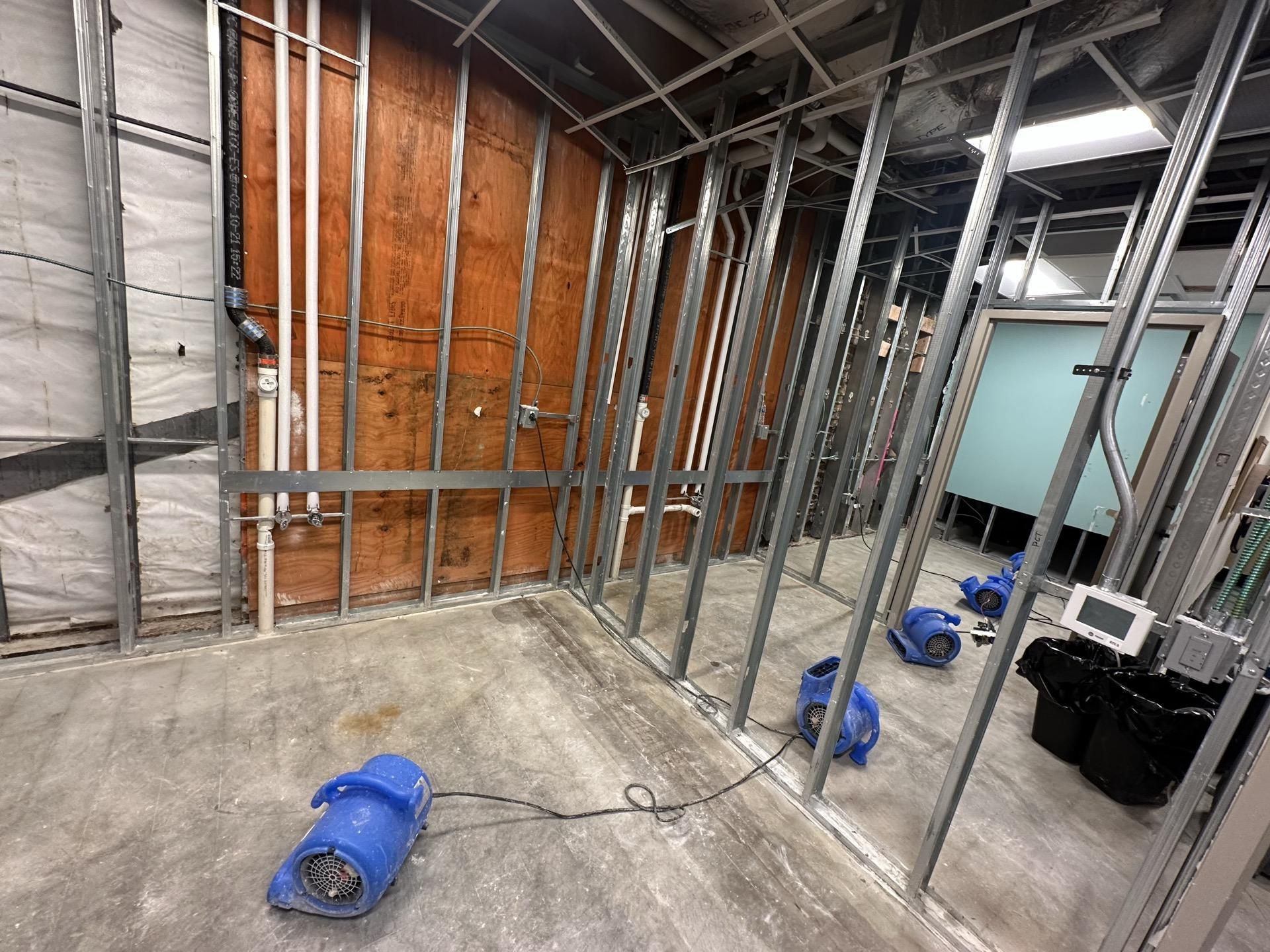 Interior of a room under renovation featuring metal wall studs, exposed pipes, and several blue floor fans for drying.