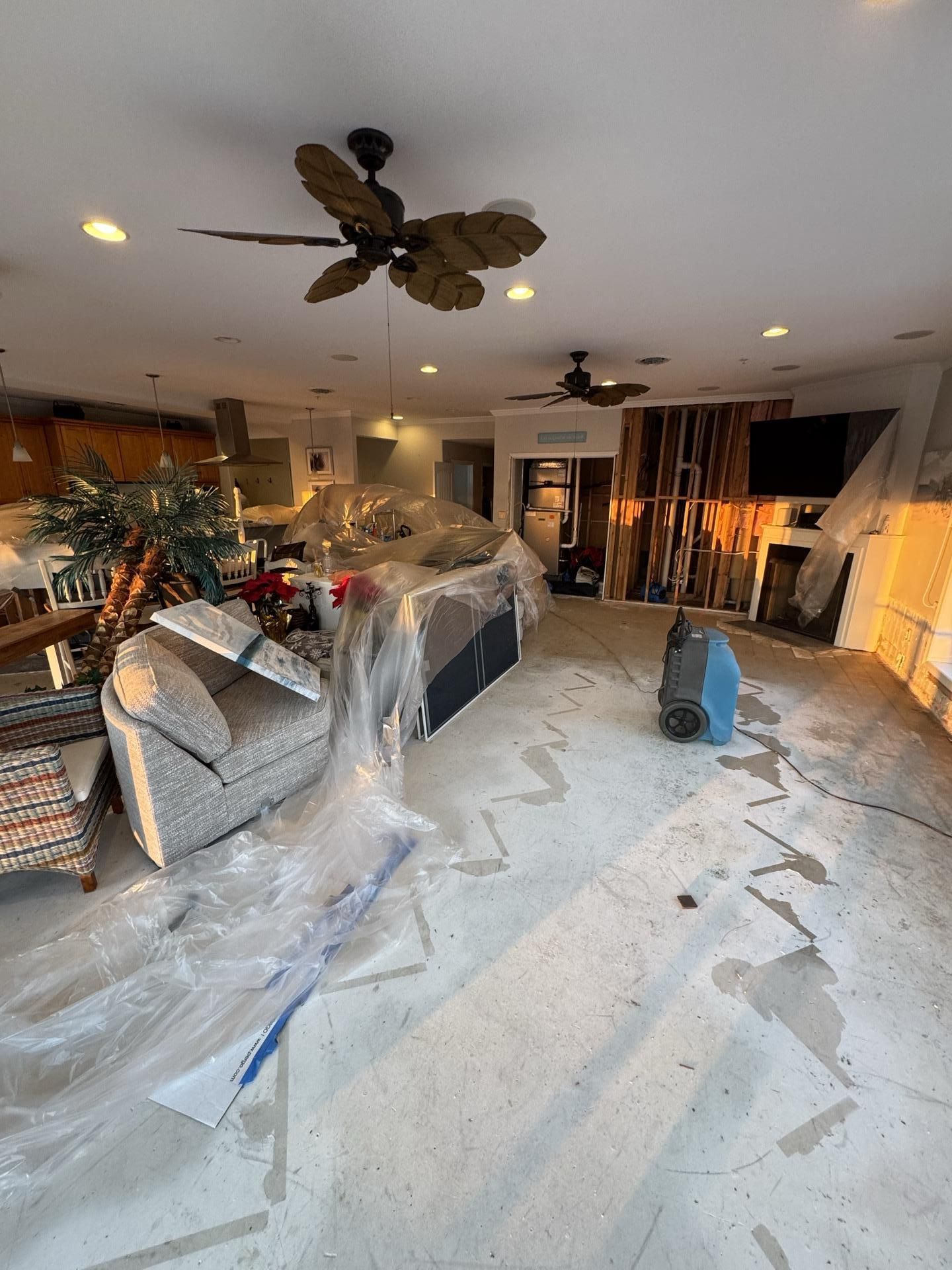 Living room under renovation with covered furniture, bare subflooring, and a portable air mover on the floor.