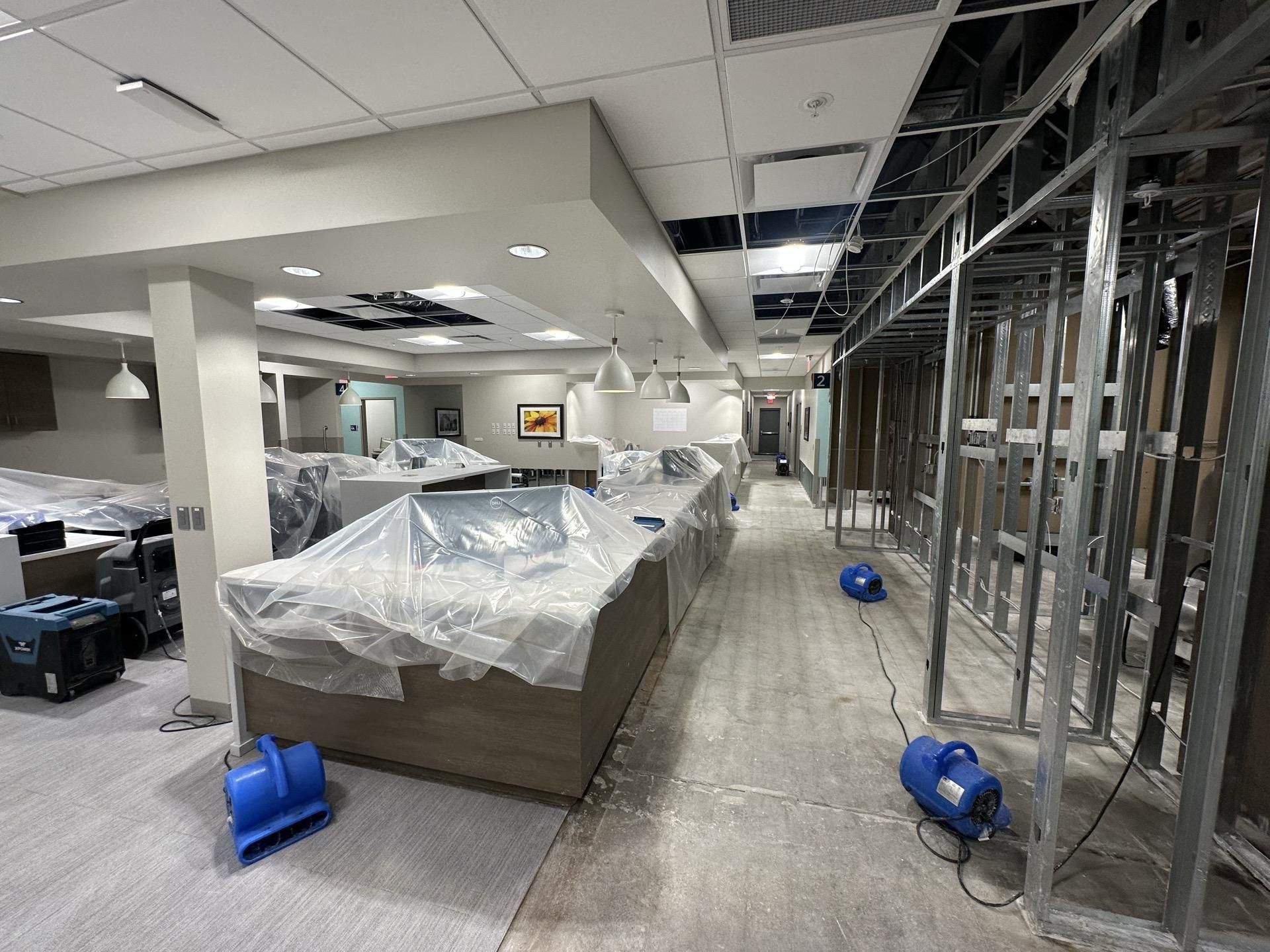 Interior construction site showing plastic-covered furniture, exposed metal wall studs, and blue air-moving fans on floor.