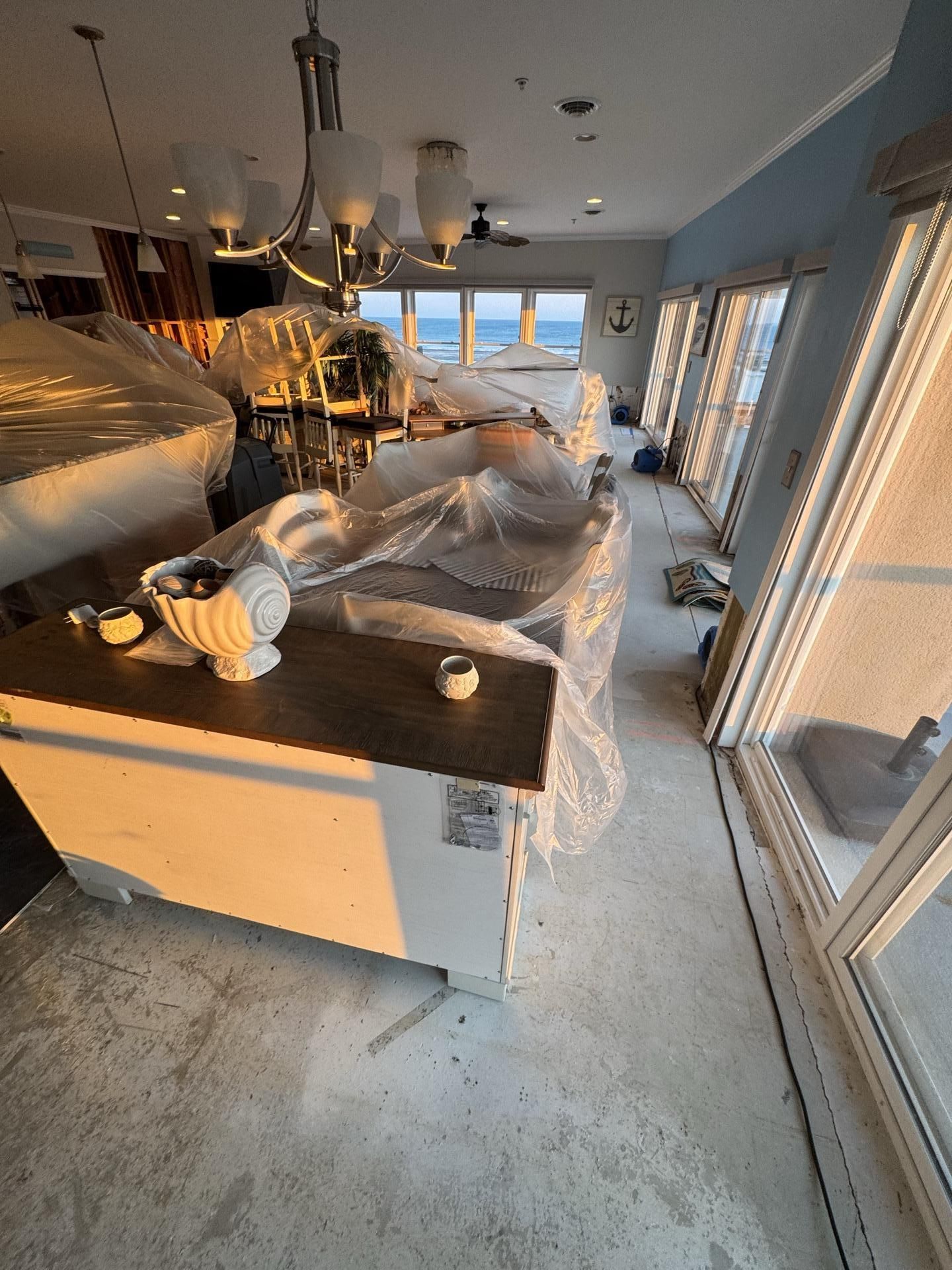 A room under renovation with covered furniture, a chandelier, and a large window overlooking the ocean.