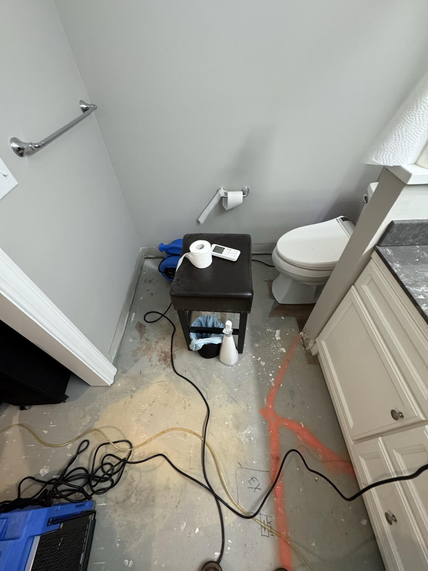 An unfinished bathroom with exposed concrete flooring, a toilet, a small stool, and electrical cords on the floor.