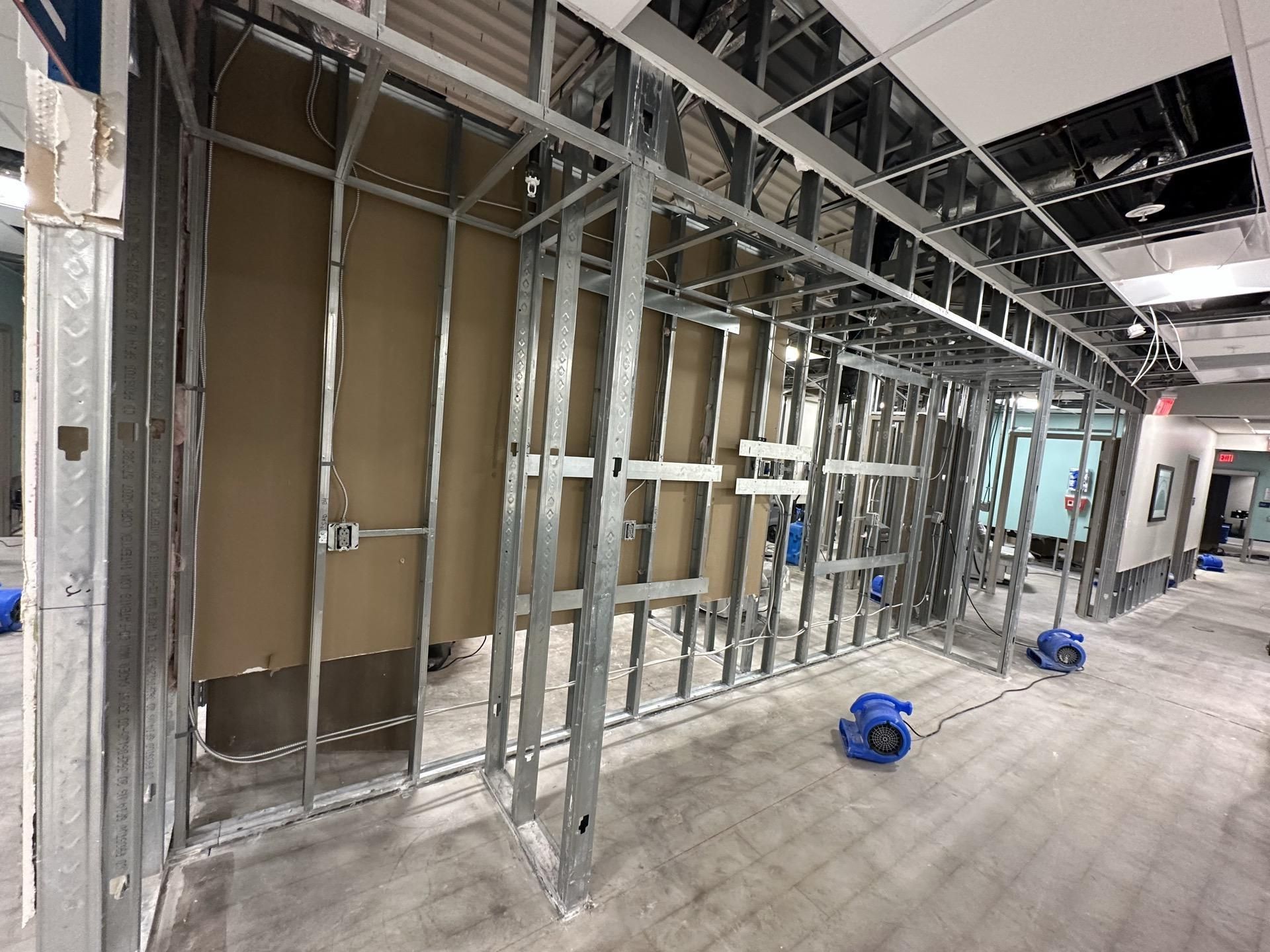 Interior of a commercial space under construction with metal wall framing, exposed wiring, and two blue floor fans.