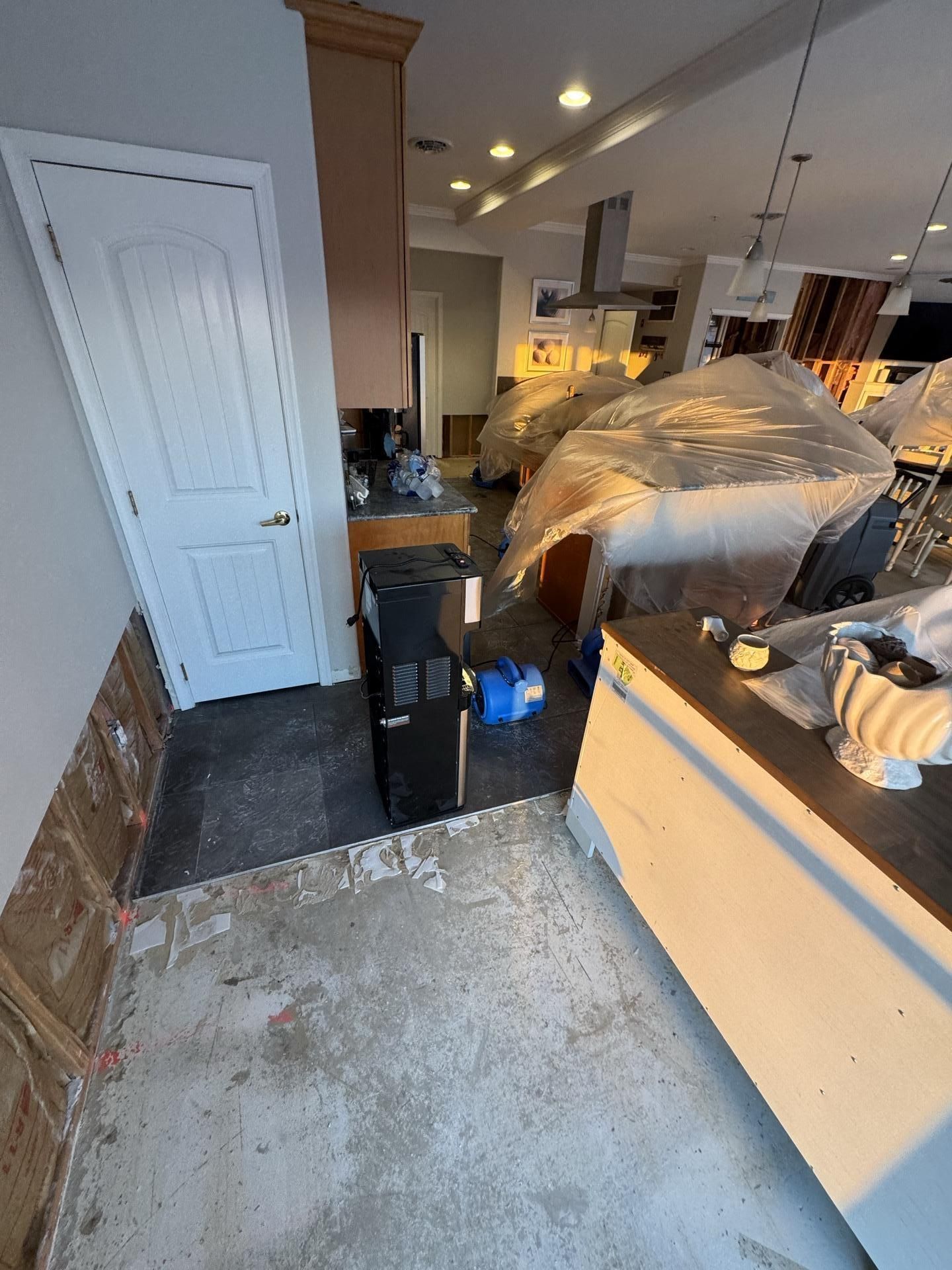 A kitchen undergoing renovation, featuring exposed subflooring, partially covered furniture, and a floor drying machine.