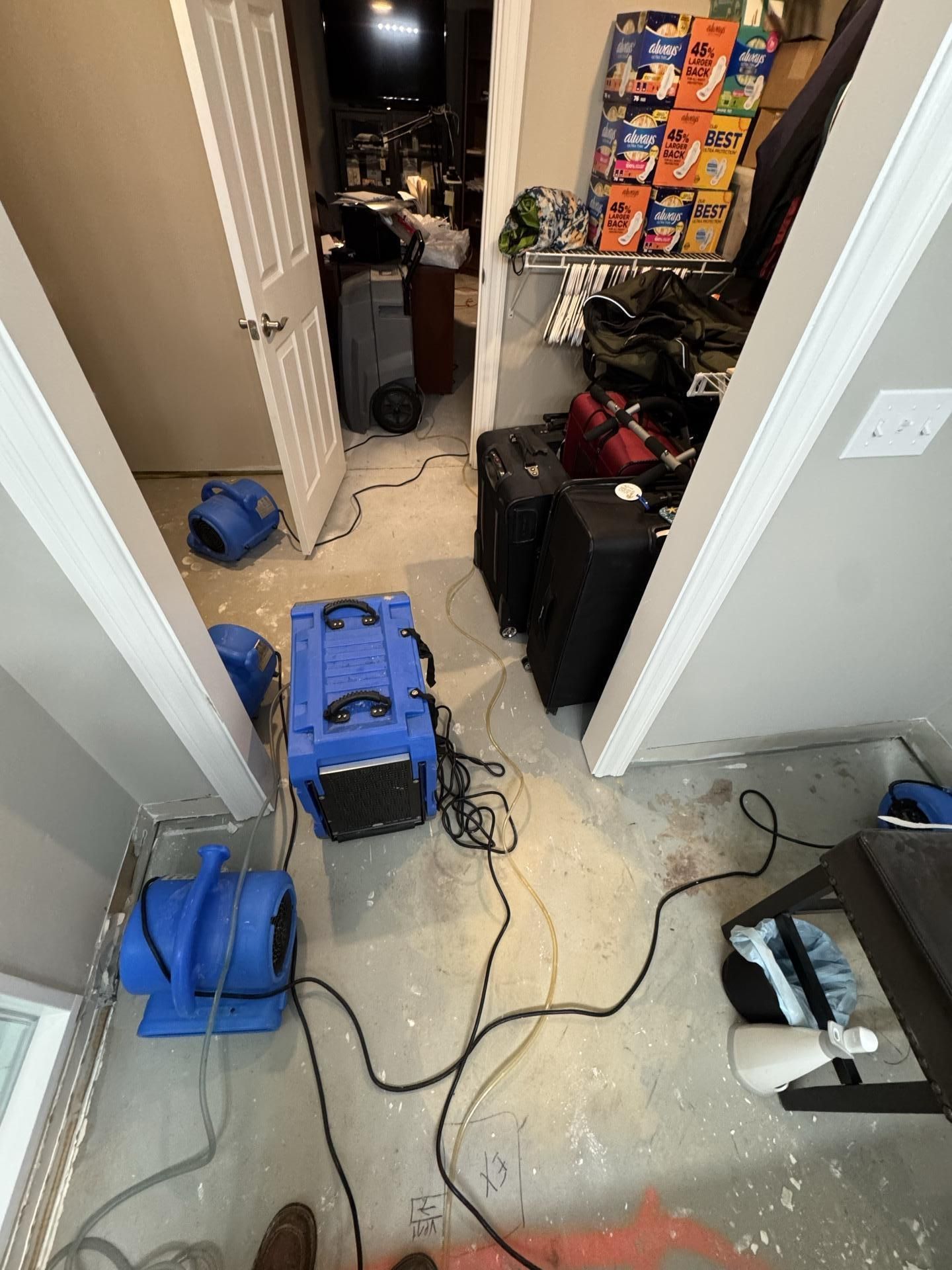 Blue industrial air movers and a dehumidifier dry a flooded room with cleared flooring, luggage, and stacked supplies.
