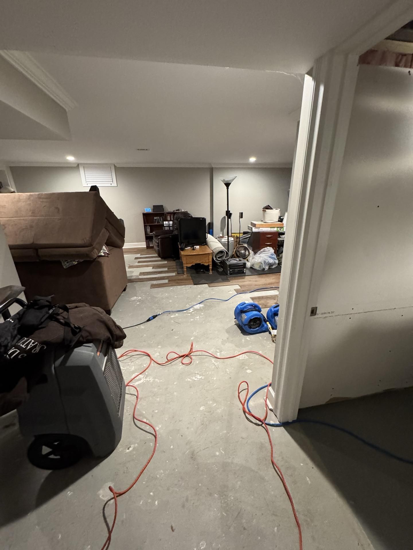 A basement under renovation with exposed concrete floors, power tools, extension cords, and furniture in the background.