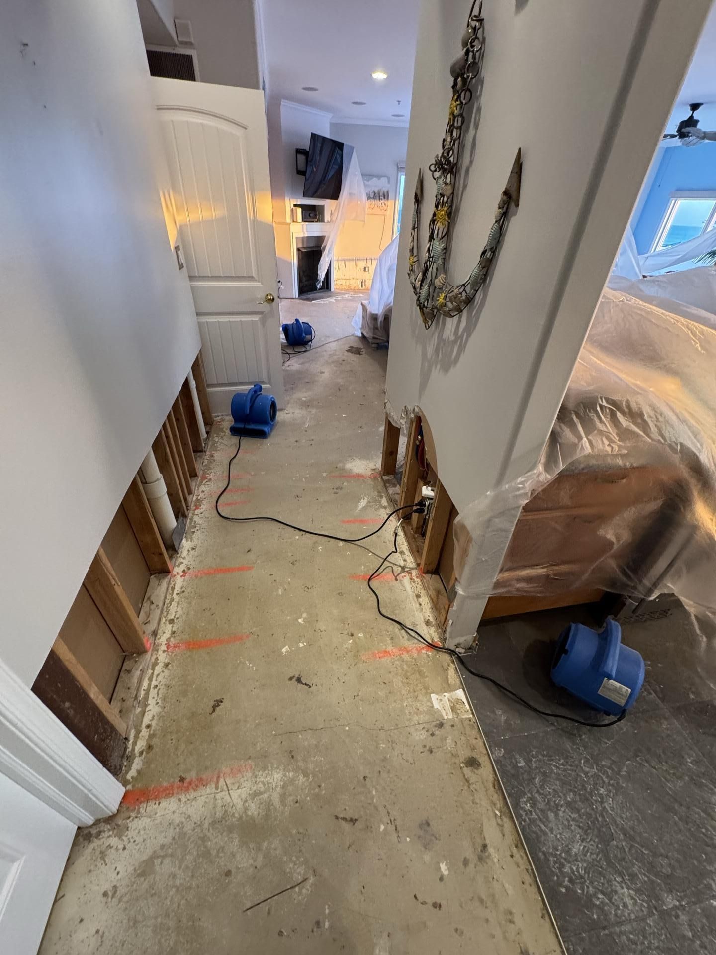 A flooded hallway with stripped walls, exposed wooden studs, and three blue industrial drying fans on the concrete floor.