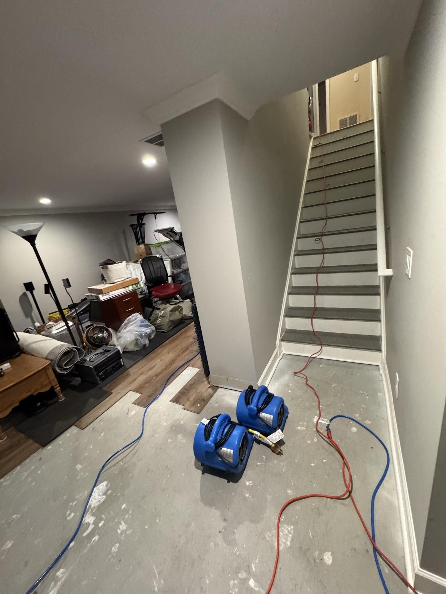 An indoor space under renovation, with exposed floor, two blue air movers, and stairs leading to an upper level.