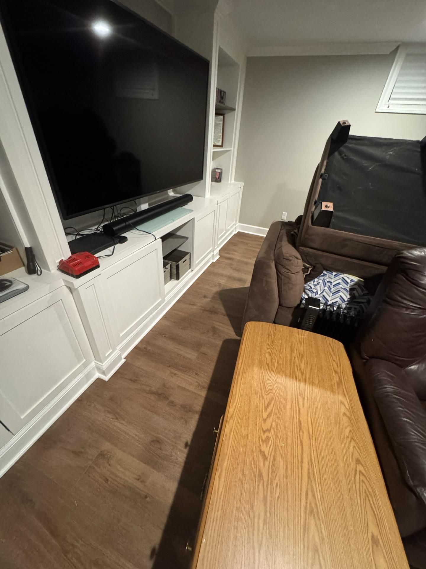 A large television mounted above white cabinets with built-in shelving, next to a brown leather sofa in a basement room.