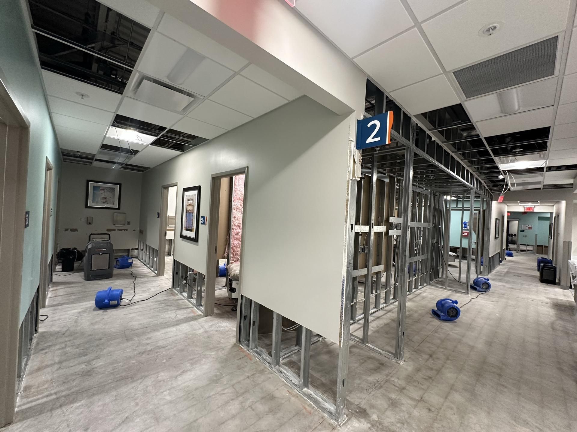 An office hallway undergoing repairs, featuring exposed metal wall studs, missing ceiling tiles, and floor drying equipment.