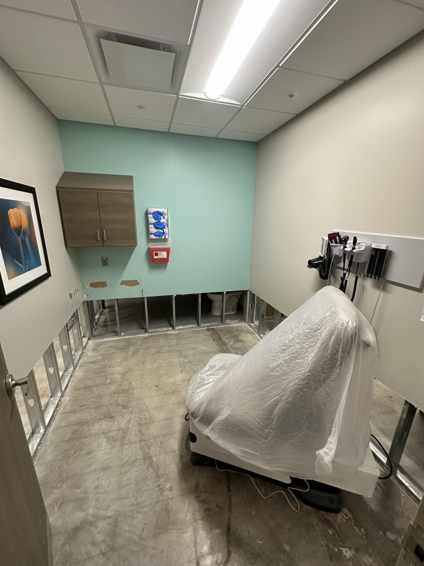 A medical exam room under renovation with exposed wall studs, a covered exam chair, and a cabinet on a mint-green wall.
