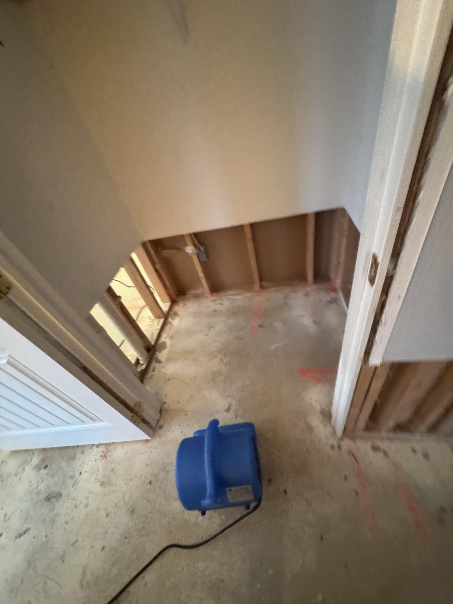 A blue air mover sits on a concrete floor in a room with partially removed drywall and exposed wall studs.