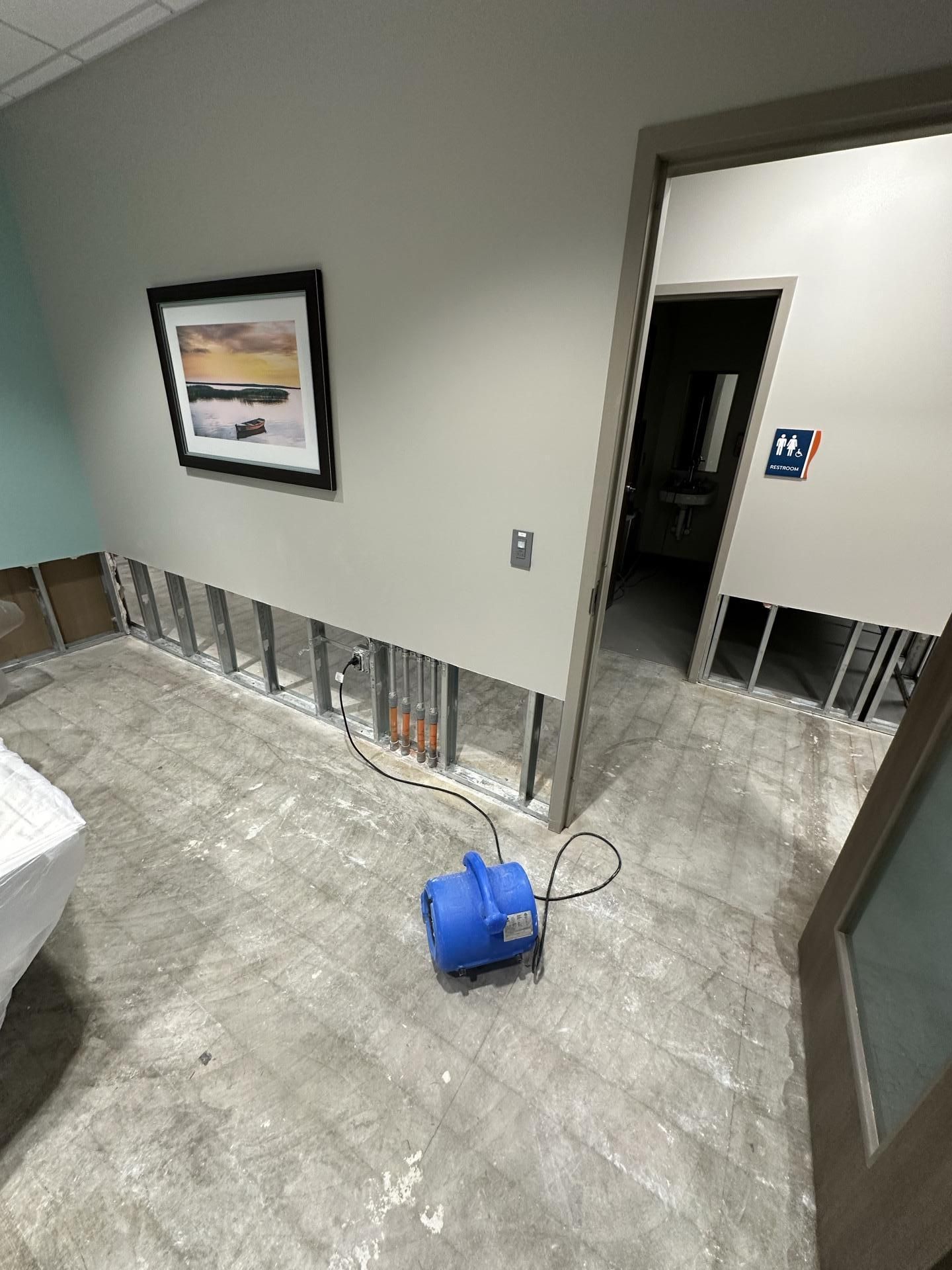 A room undergoing construction with exposed wall studs, a gray floor, a framed landscape photo, and a blue air blower.
