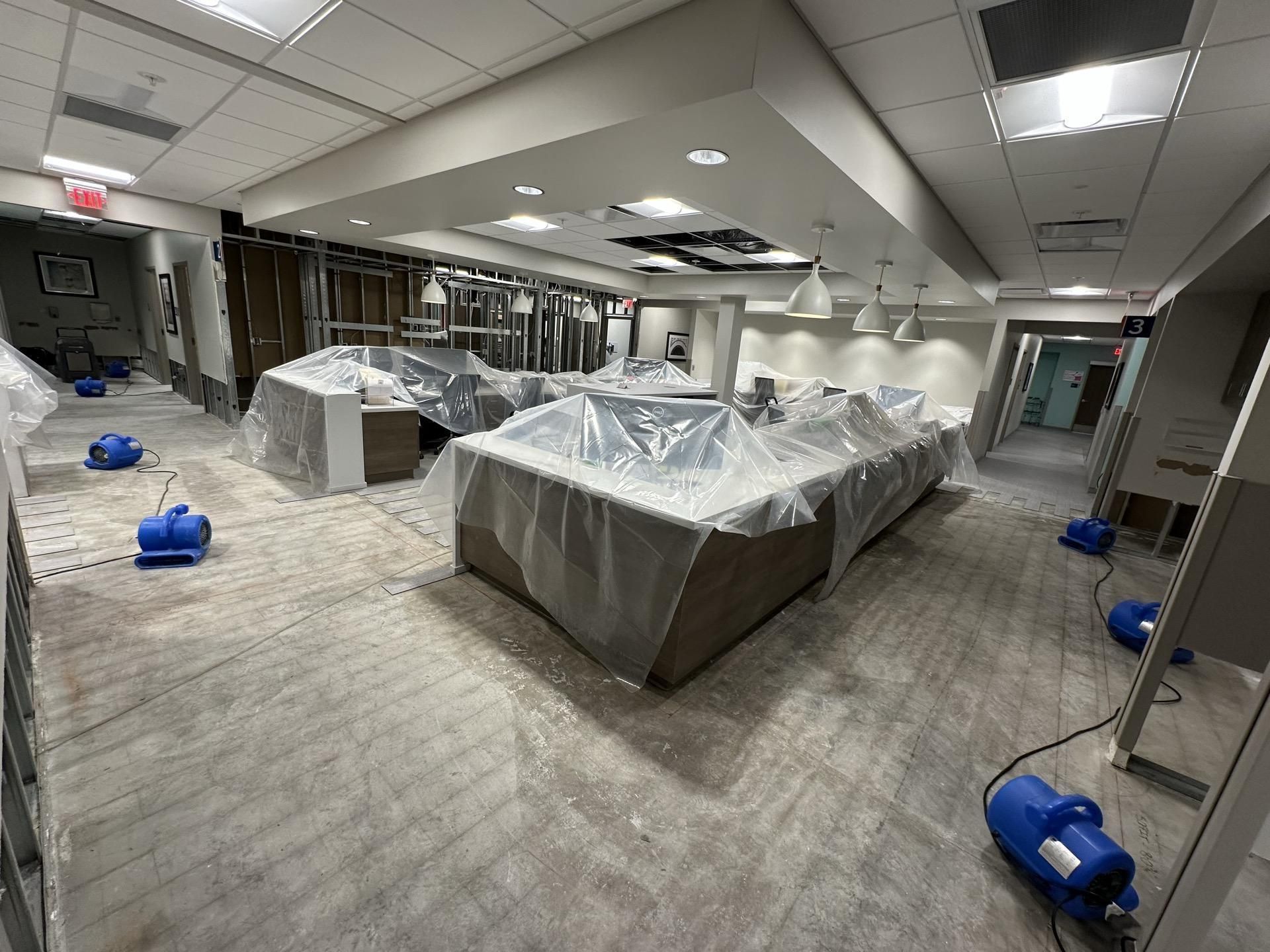A construction site in an office with furniture covered in plastic sheeting and several blue air movers on the floor.