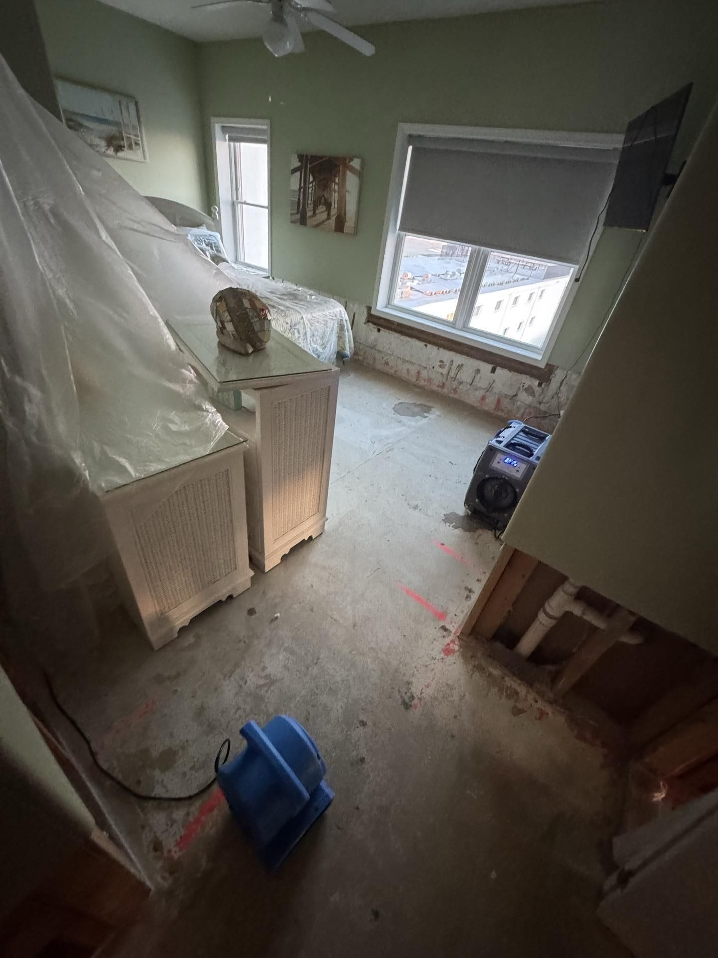 A room under renovation with exposed floor sub-base, plastic-covered furniture, and industrial air-drying equipment.