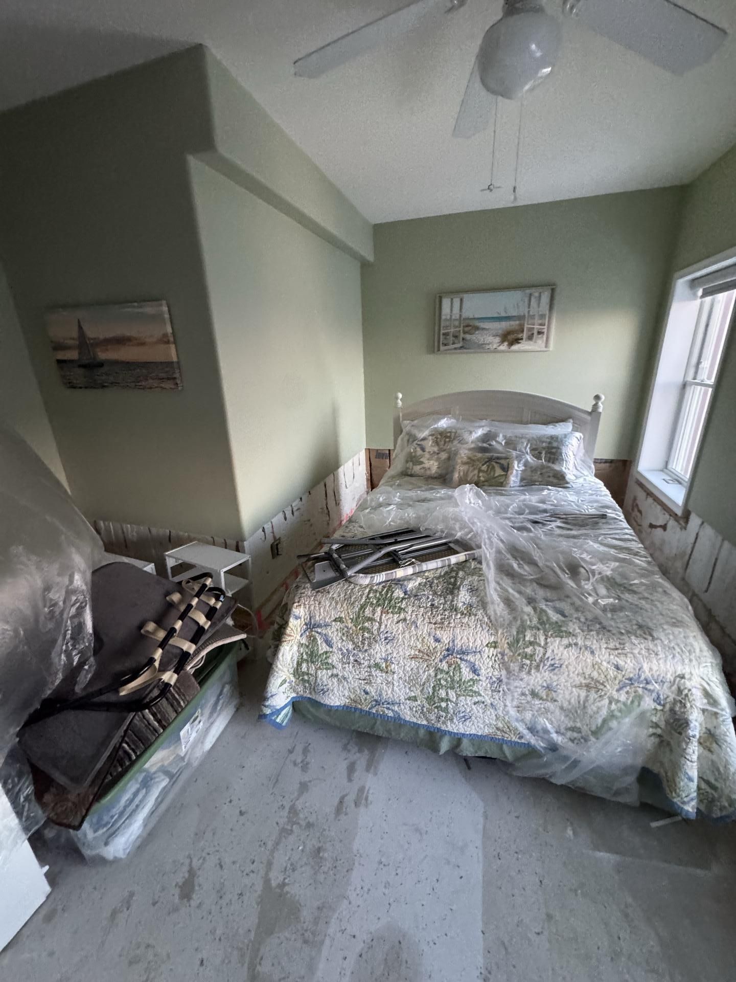 A bedroom with a bed and a plastic-covered storage bin, featuring light green walls, a ceiling fan, and framed art.