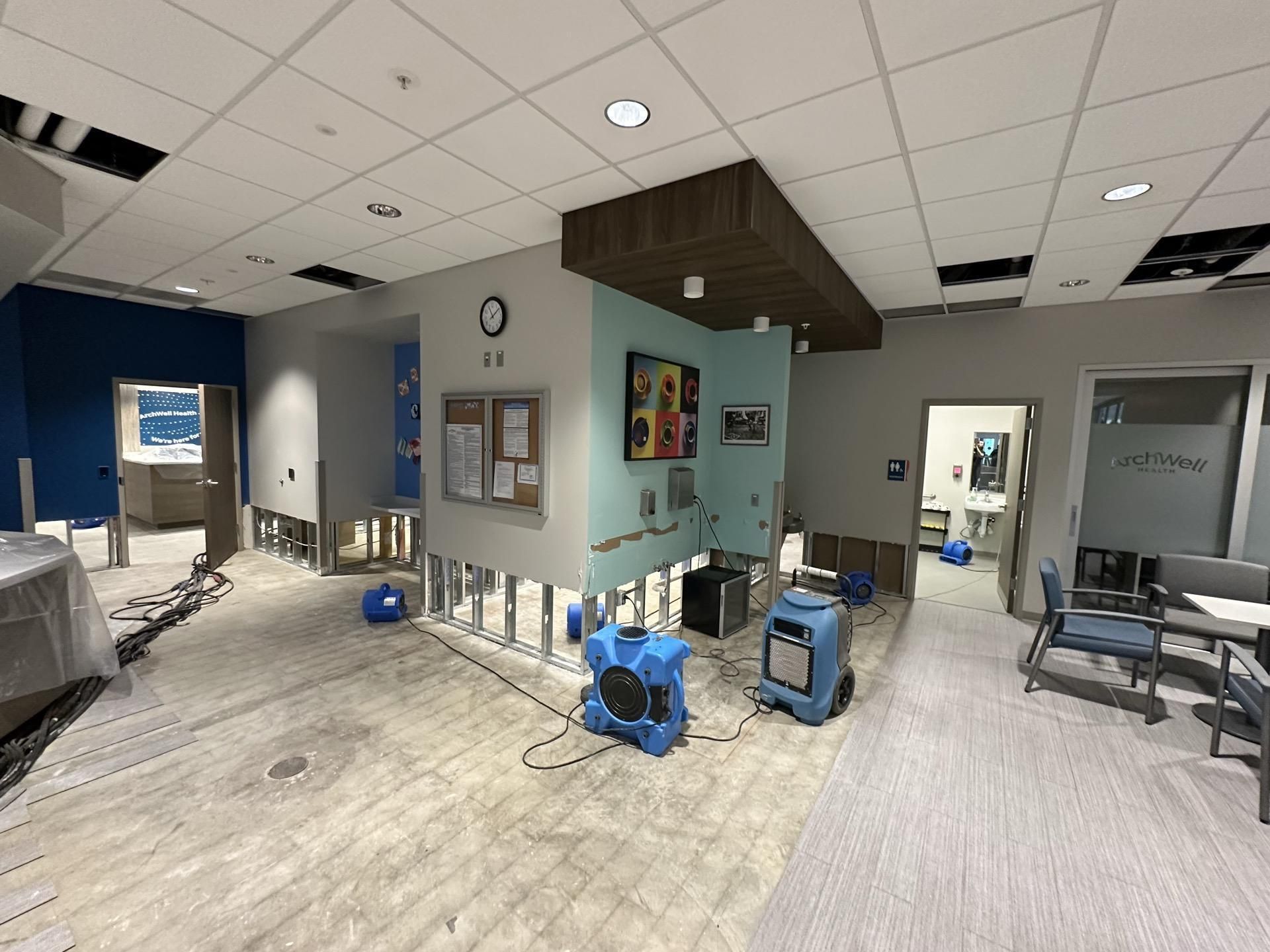 Office interior under restoration with exposed wall studs, blue drying equipment on the floor, and missing ceiling tiles.