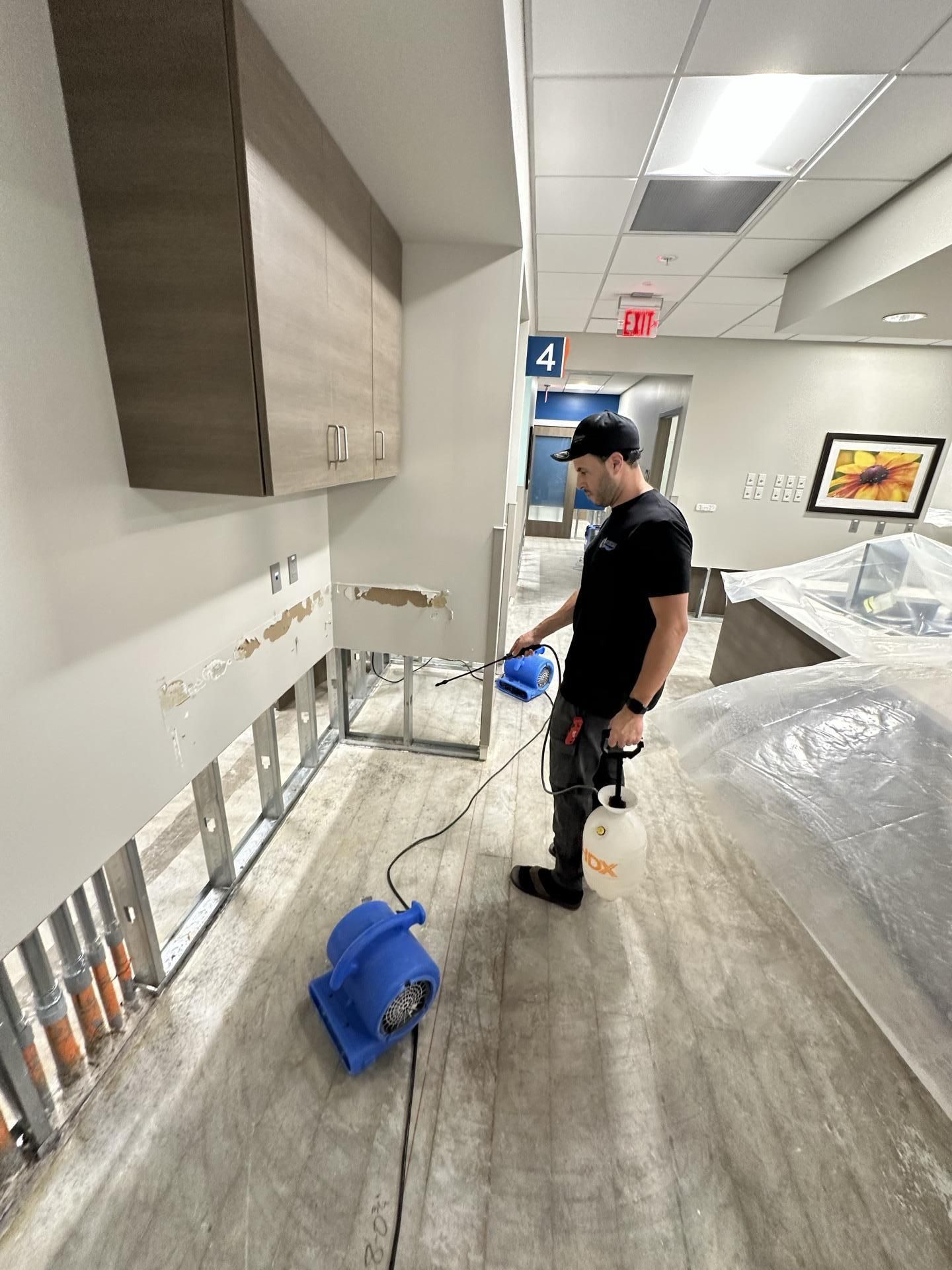 A professional in a black shirt cleans a wall area with a spray bottle and blue air mover in a room under renovation.