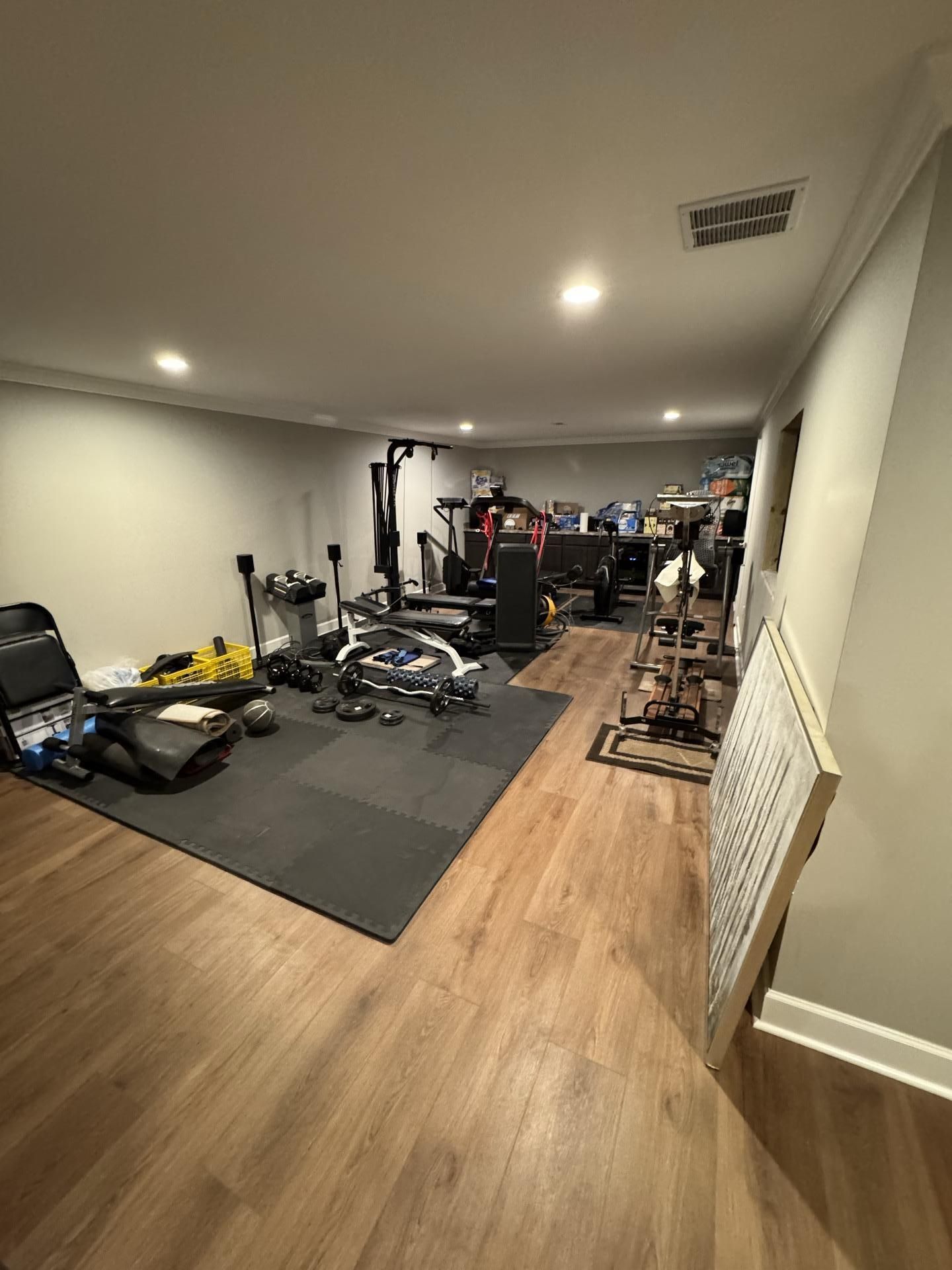 A home gym setup with workout equipment, including a weight bench and stationary bike, on a grey mat in a basement room.