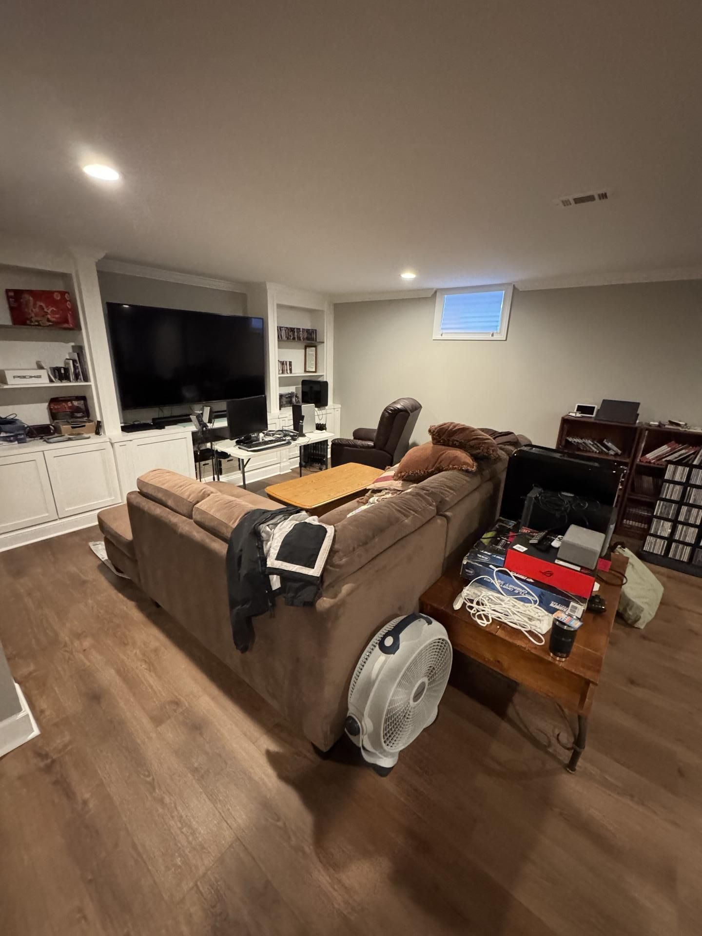 A cozy, modern basement living room with a brown sectional couch, a large TV, built-in shelving, and a small window.