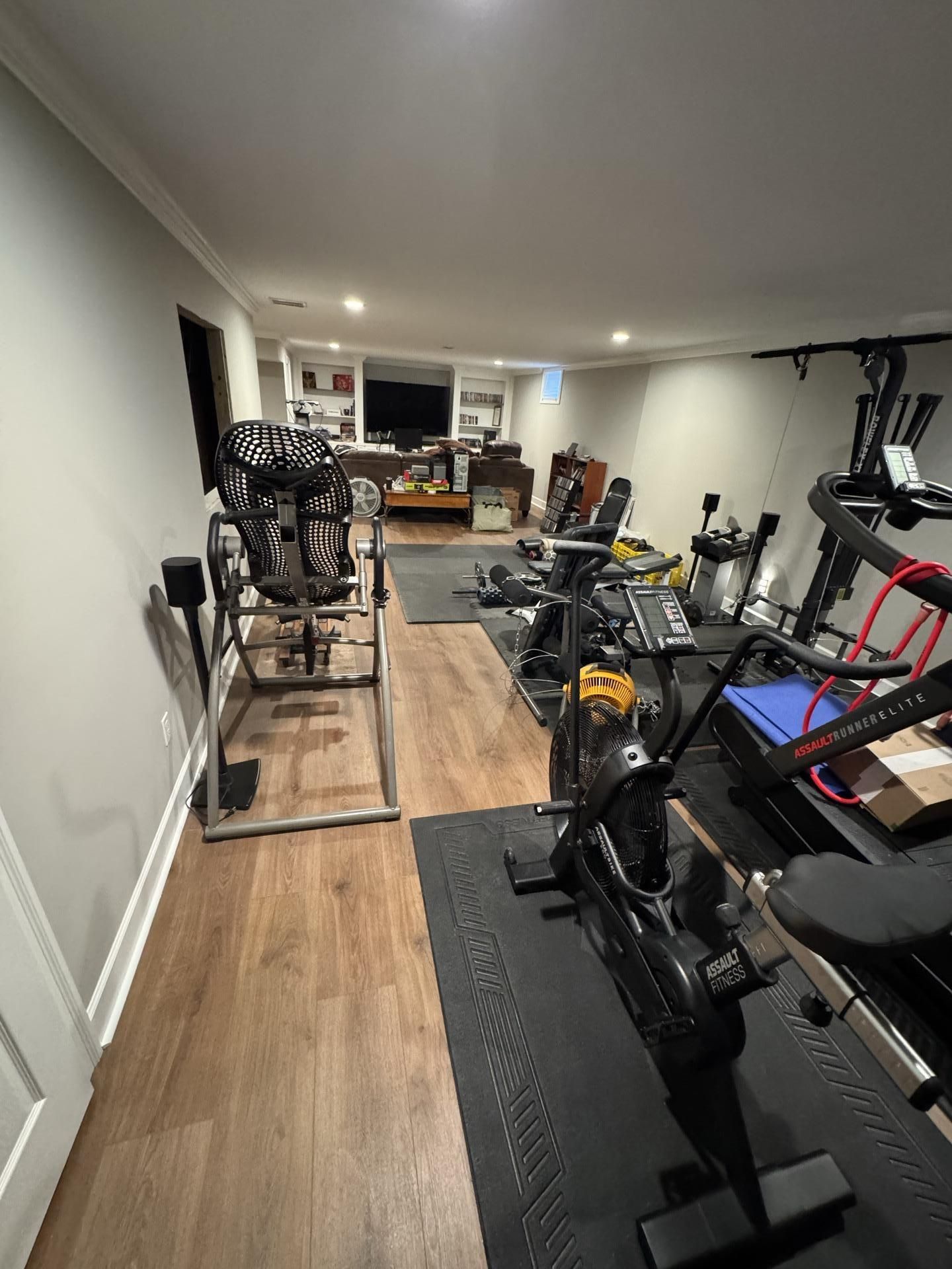 A home gym basement with workout equipment including a rowing machine, inversion table, and treadmills on wood floors.