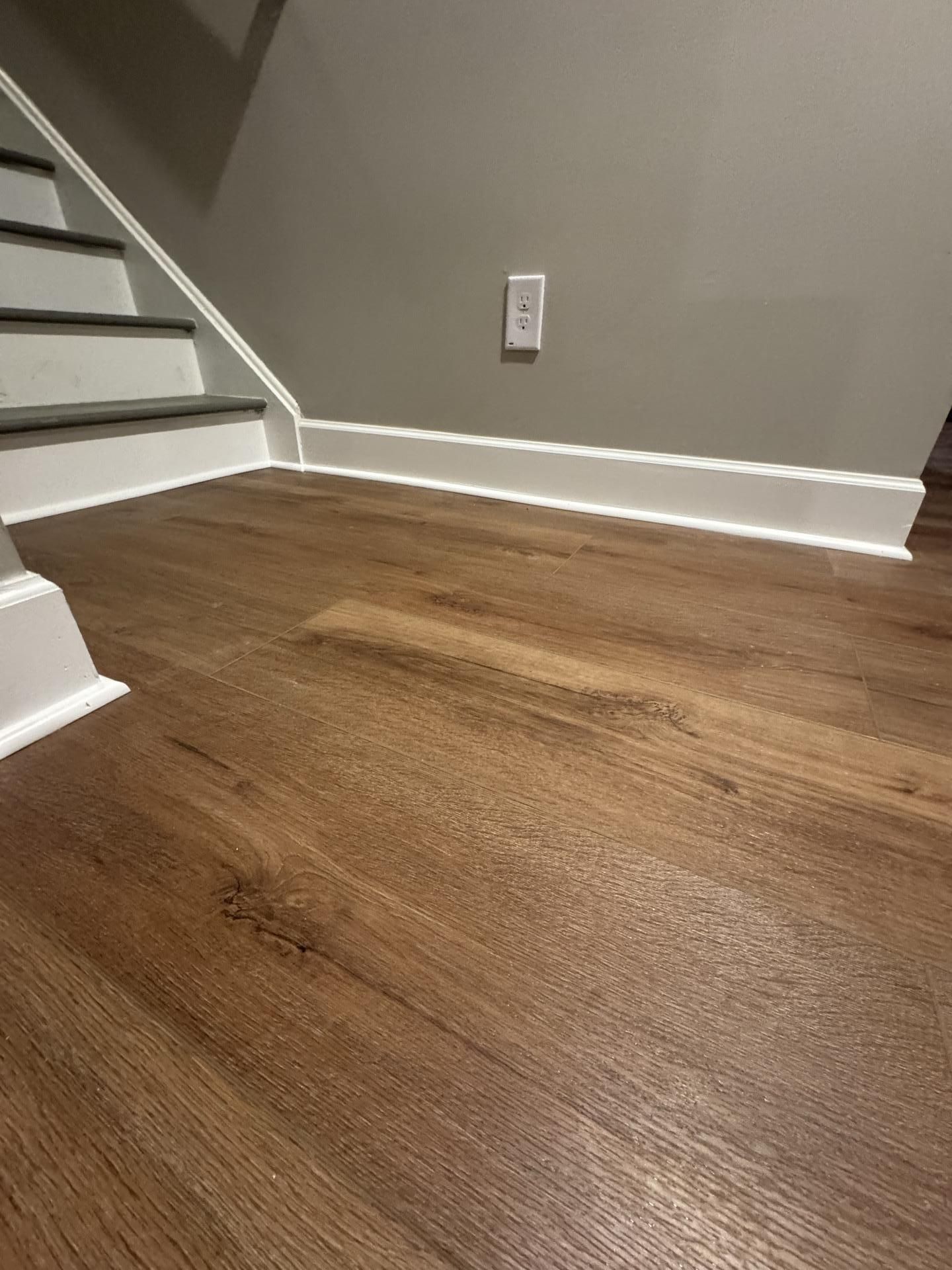 A low-angle view of wood-look flooring next to white baseboards and the bottom of a staircase in a gray-walled room.