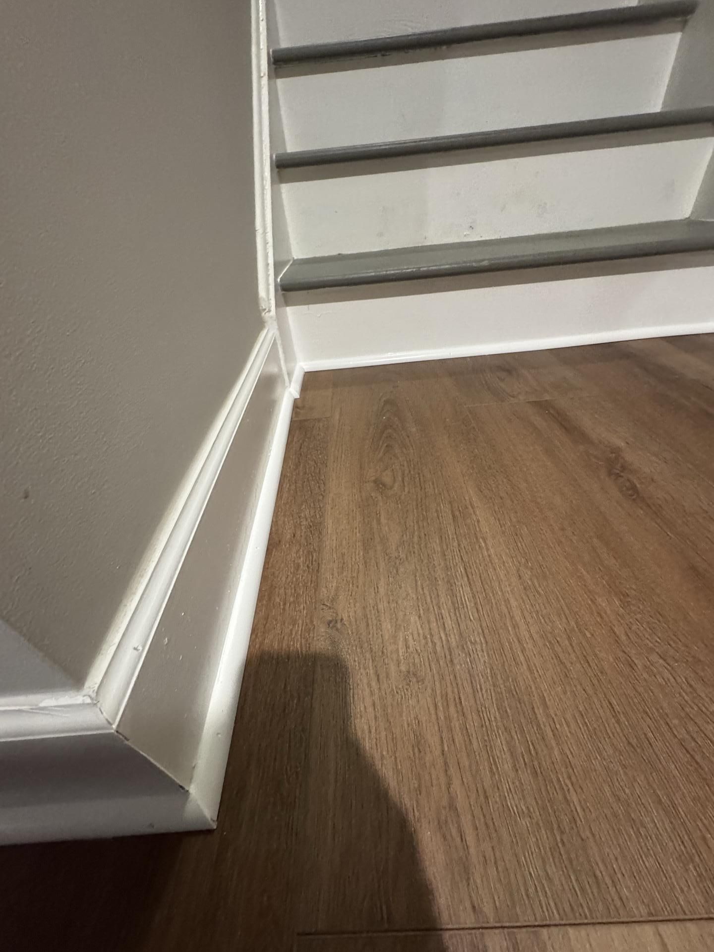 A low-angle view of a white baseboard meeting a light brown wood floor next to the bottom of a staircase.