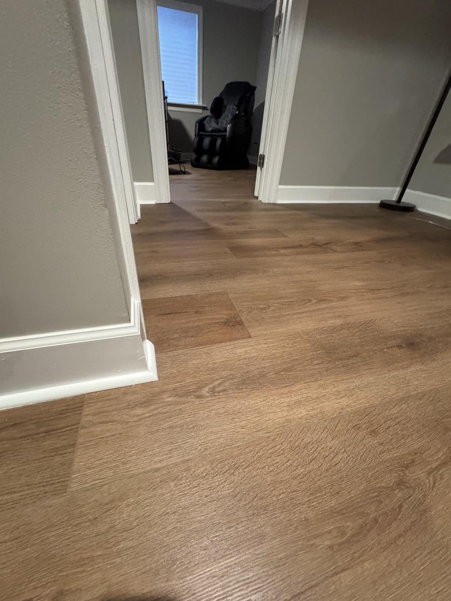 A view of a modern hallway with light brown wood-look flooring, gray walls, white trim, and a doorway leading to a room.