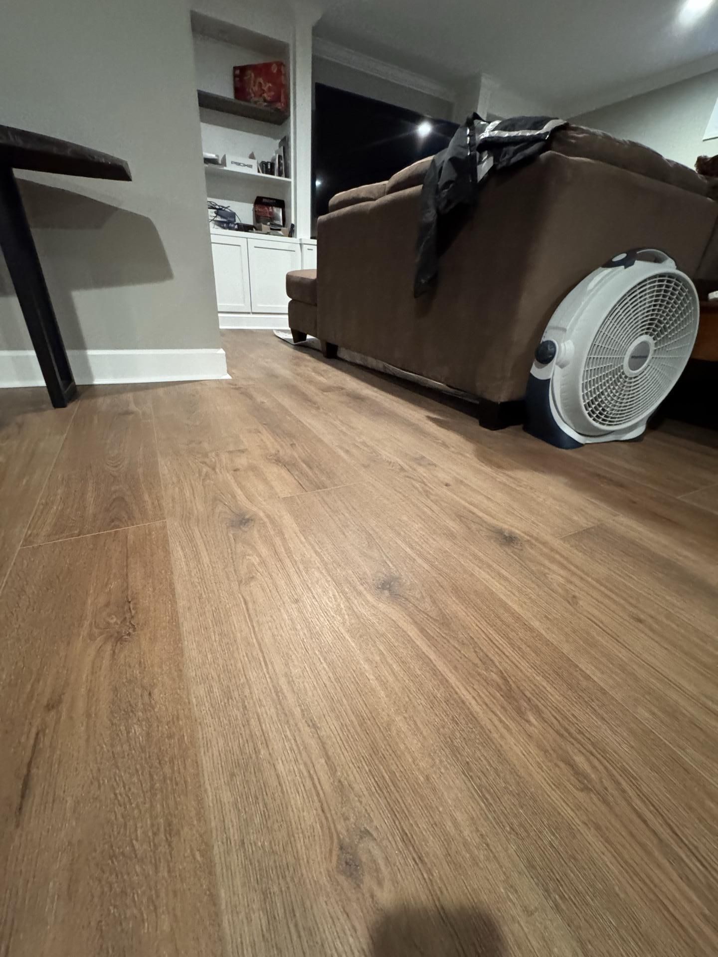 Low-angle view of wood-look plank flooring in a living room with a brown sofa and a floor fan nearby.