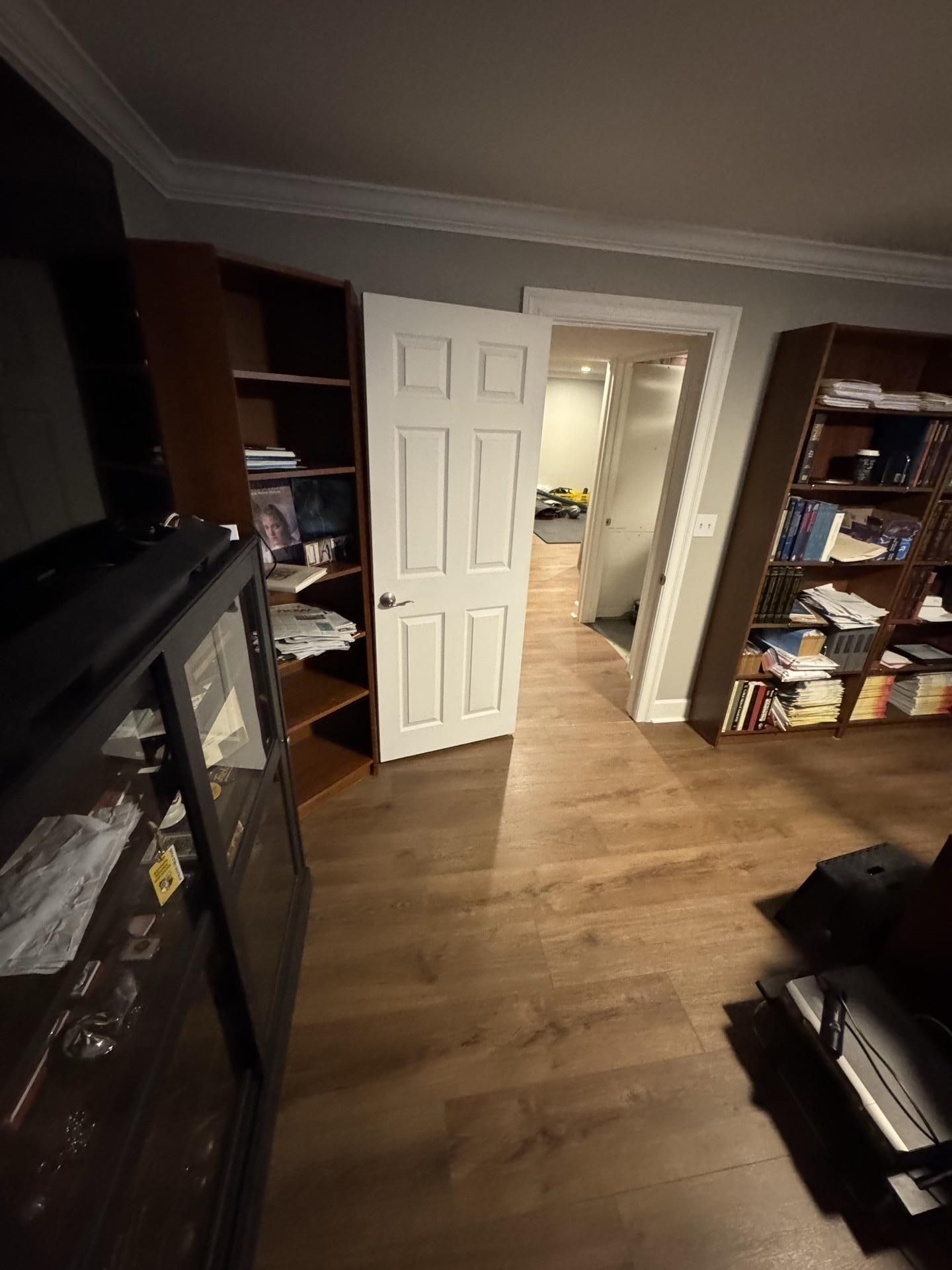 A wide-angle view of a room with light wood floors, two wooden bookcases, a glass-front cabinet, and an open white door.