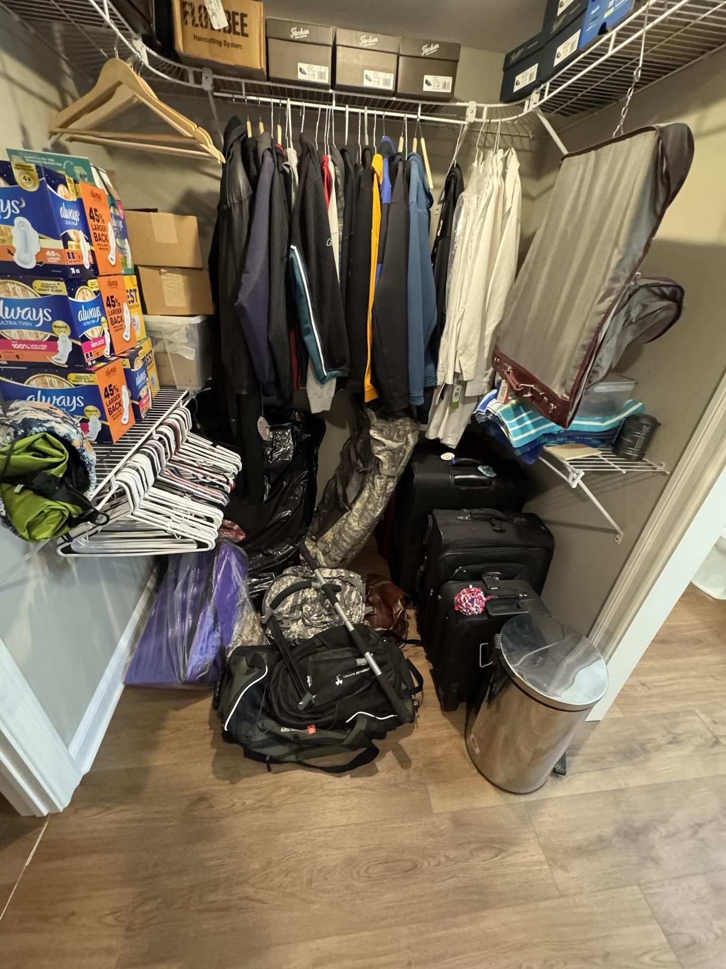 A cluttered walk-in closet with clothes hanging on a rack, stacked boxes, luggage on the floor, and a metal trash can.