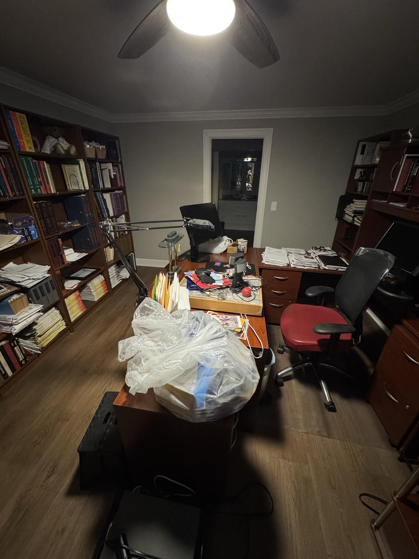 A home office featuring a desk piled with clutter, rolling chairs, and dark wood bookshelves filled with books.