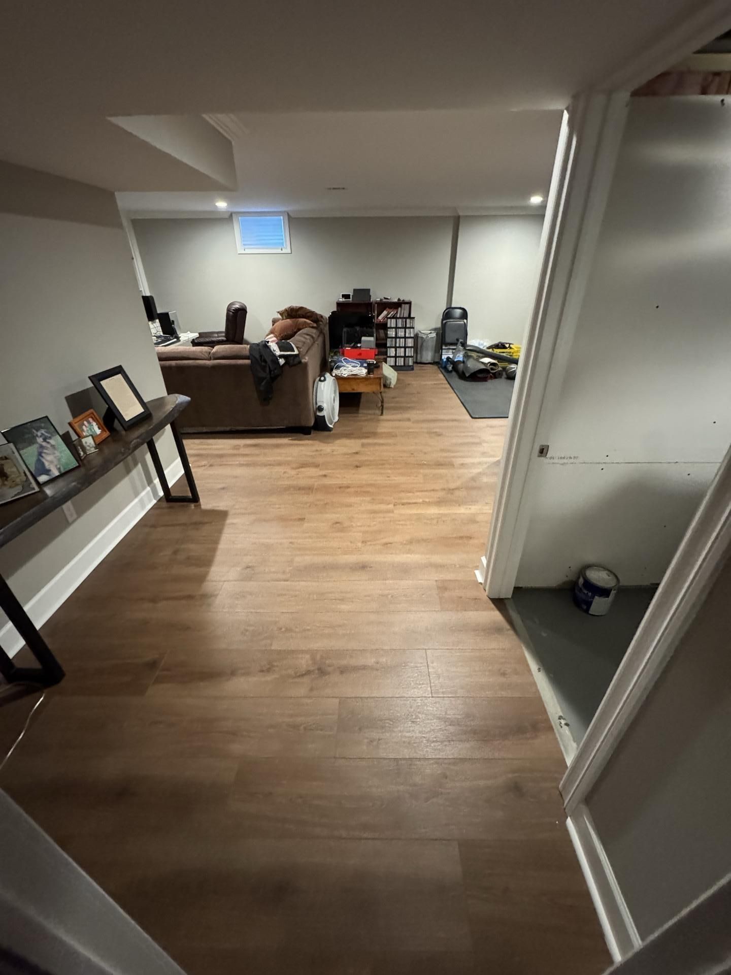 A wide-angle view of a finished basement with light wood-tone flooring, neutral walls, and furniture in the background.