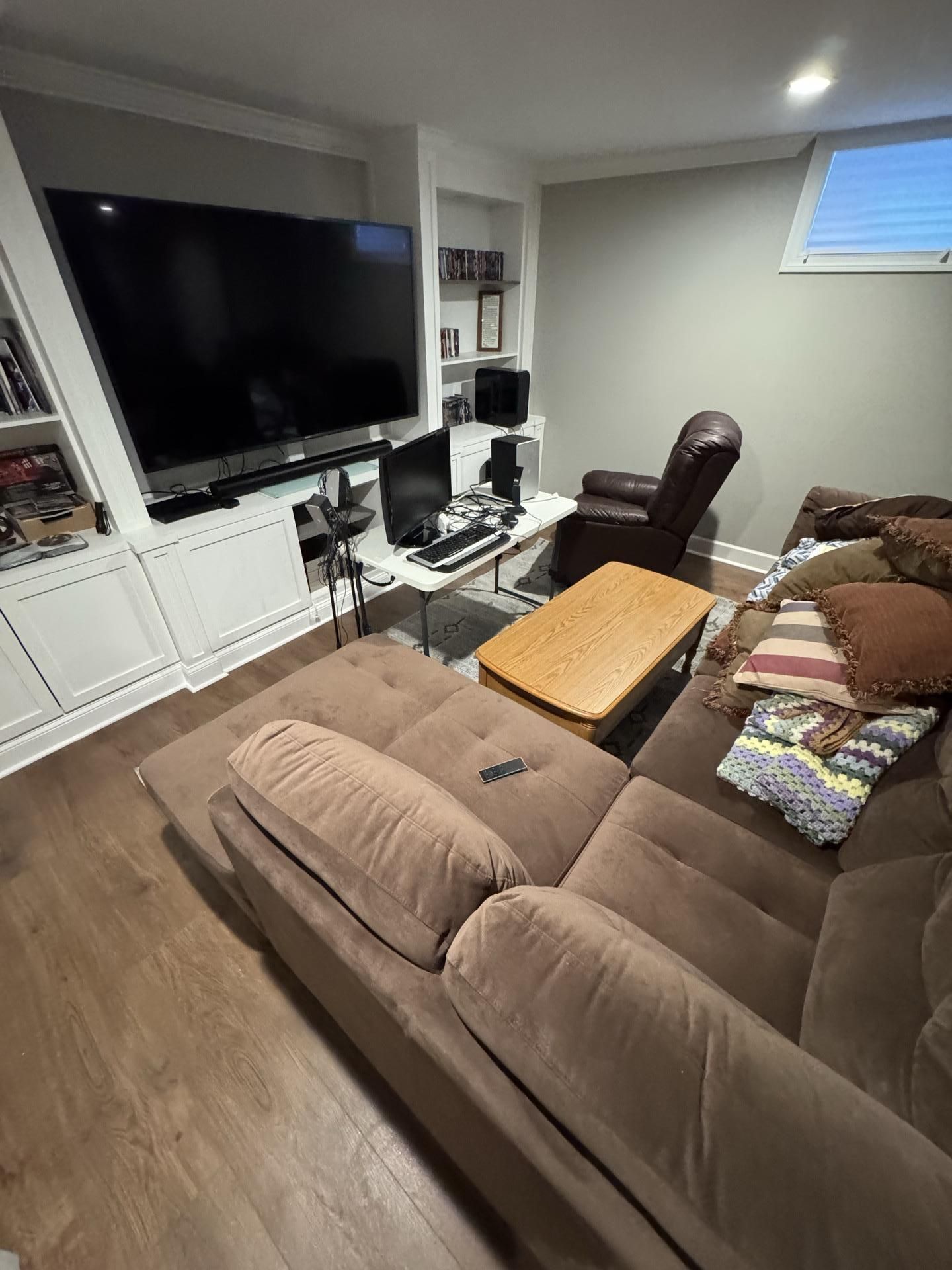 A living room with a large TV, white built-in shelving, a brown sectional sofa, a coffee table, and a brown recliner.