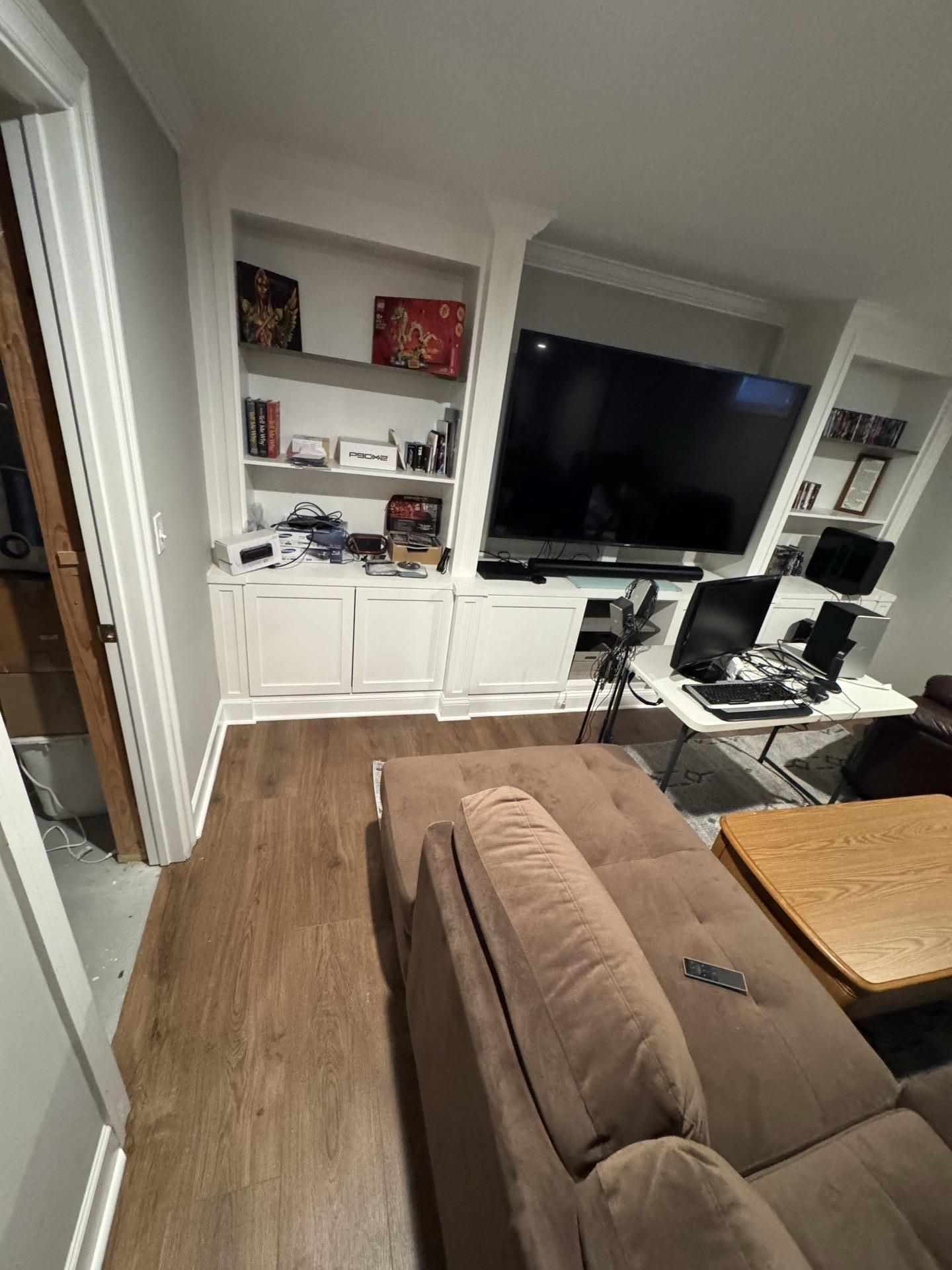 A living room featuring white built-in shelves, a large television, a brown sofa, and a coffee table.