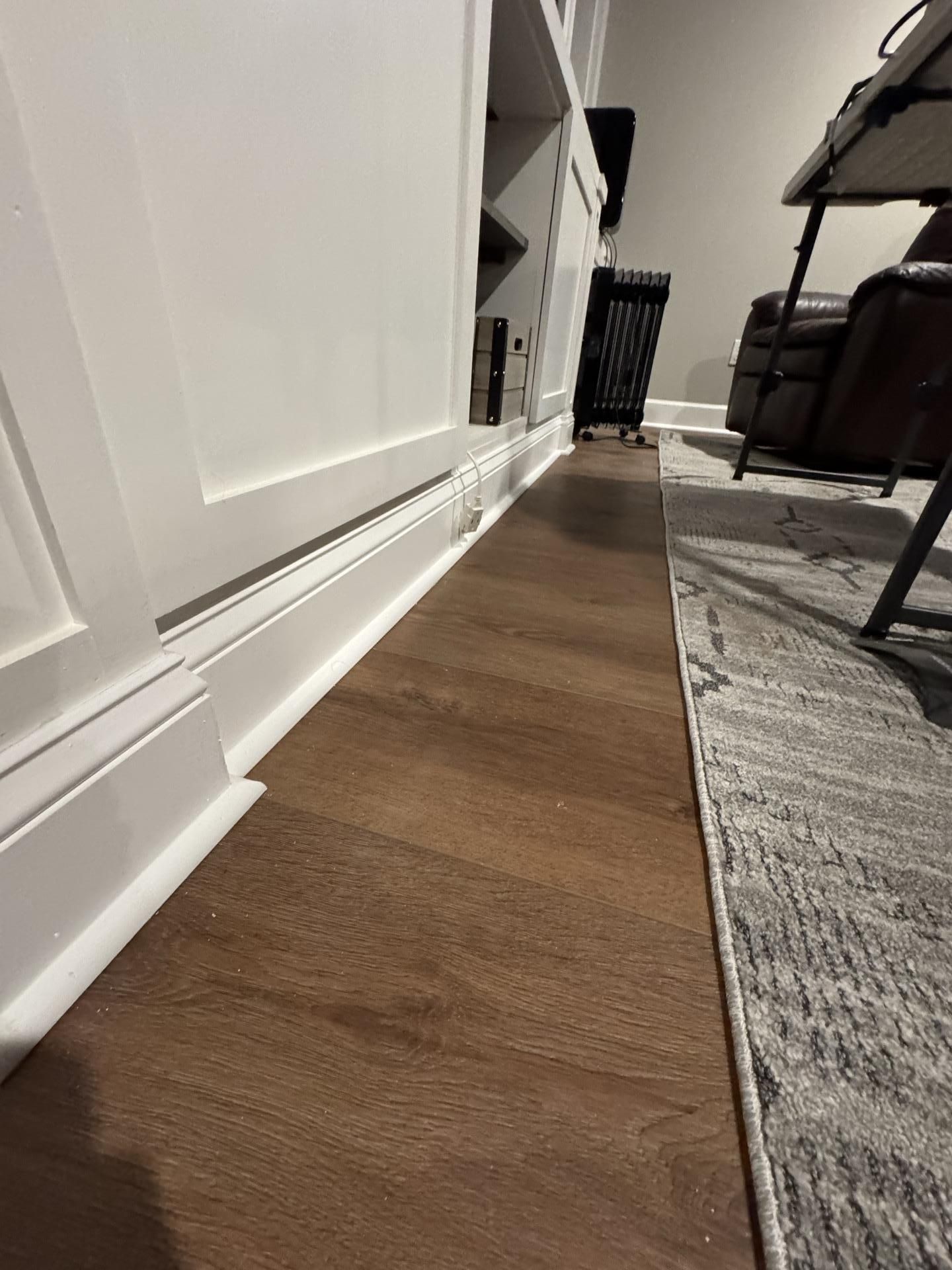 A floor-level view of a wood-look floor meeting a white baseboard cabinet, with a grey patterned rug on the right.