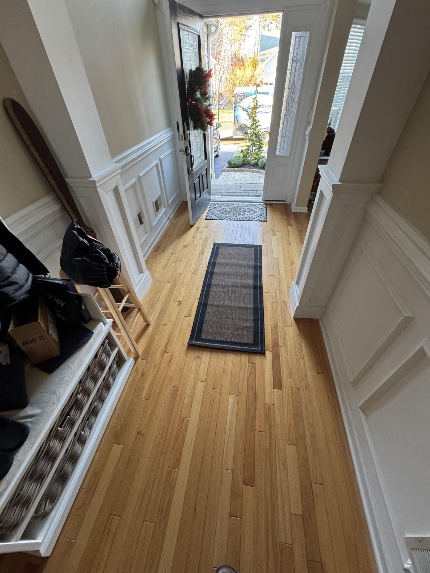 A light-filled entryway with wood floors, white wainscoting, a shoe bench, and a runner rug leading to an open front door.