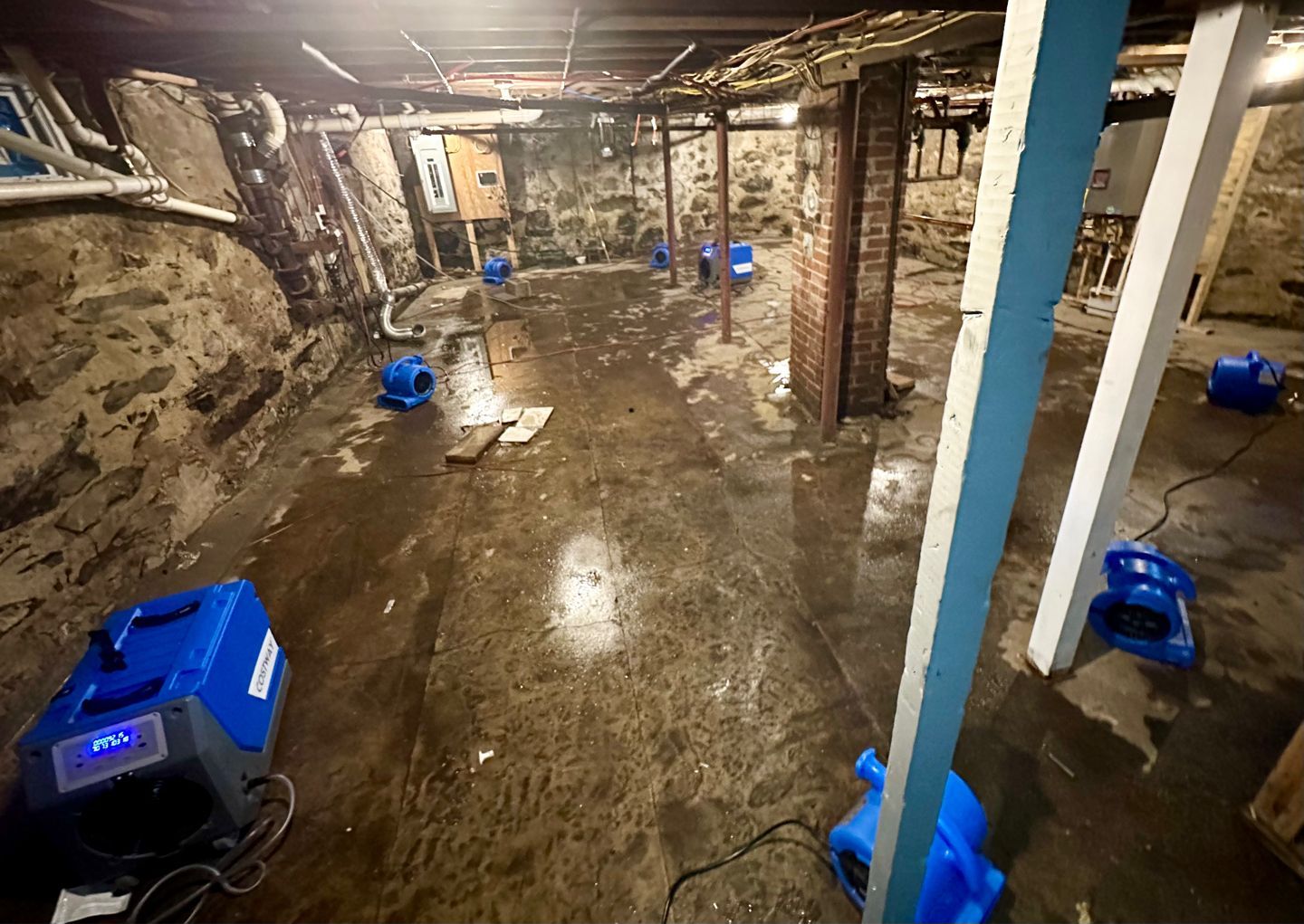 A flooded basement with wet stone walls and floor, featuring several blue industrial fans drying the area.