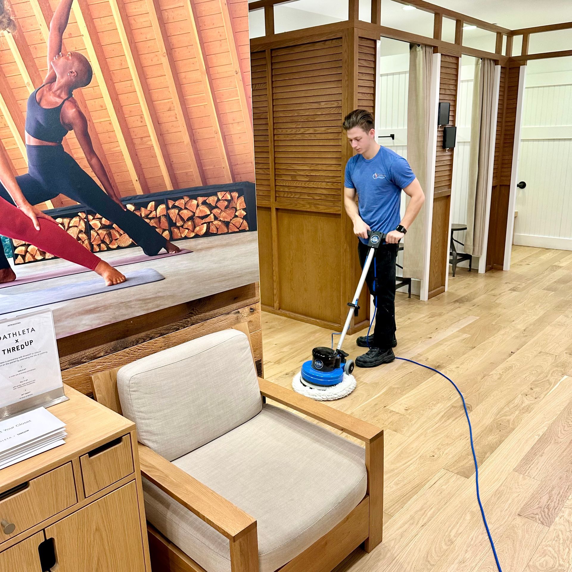 A worker in a blue shirt uses a floor buffer on wooden flooring in a yoga studio with a large mural on the wall.