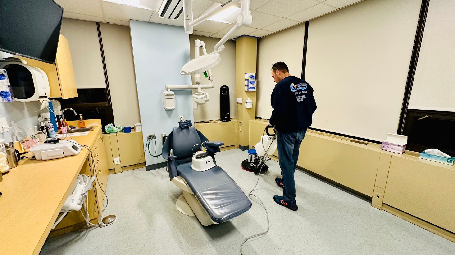 A person in a dark uniform uses a floor cleaning machine in a dental exam room with a reclining chair.