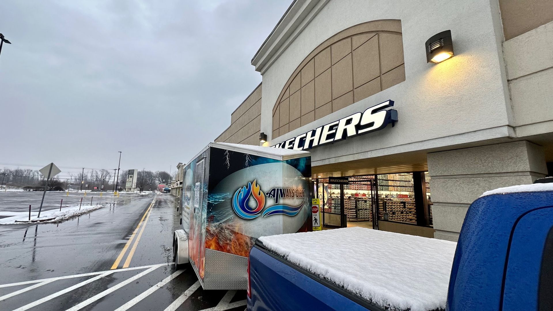 A Skechers store exterior with a truck pulling a graphic-covered trailer parked in front on a snowy day.