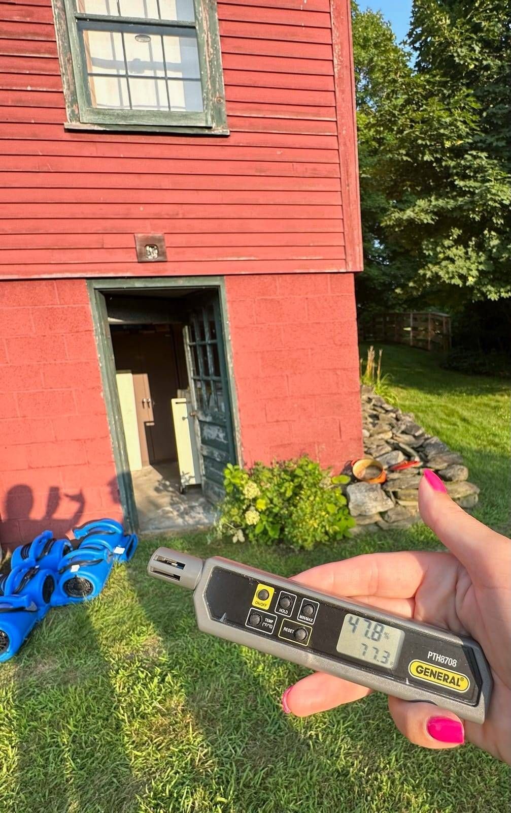 A person holds a digital moisture meter displaying 47.8% against a red building with an open door on a sunny day.