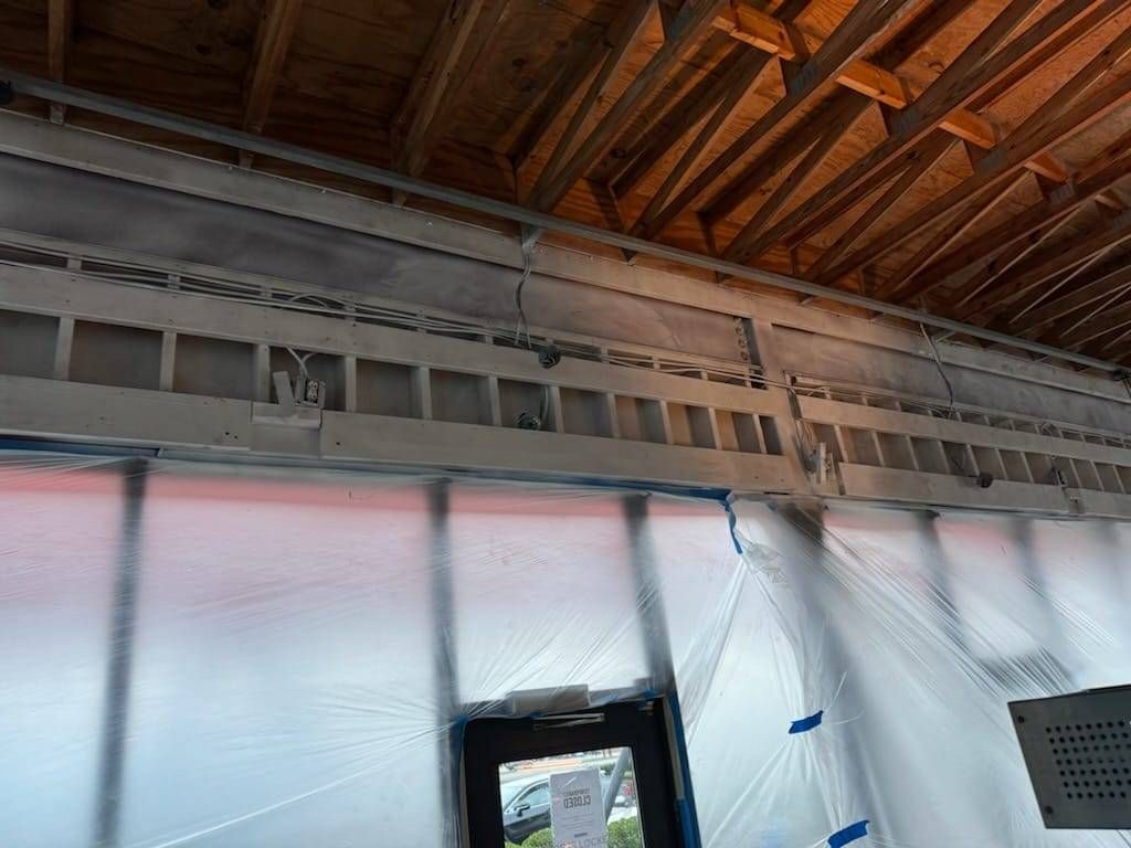 Interior view of a building under renovation, showing exposed ceiling joists above a wall frame covered in plastic sheets.