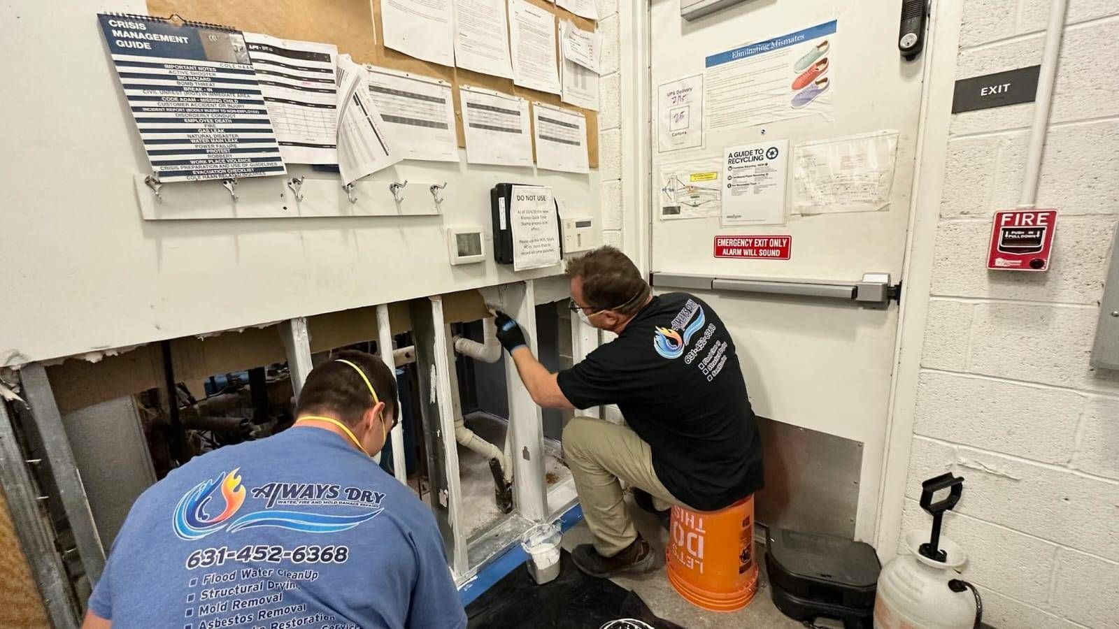 Two plumbers in branded shirts repair pipes behind a wall section in an office or utility room.