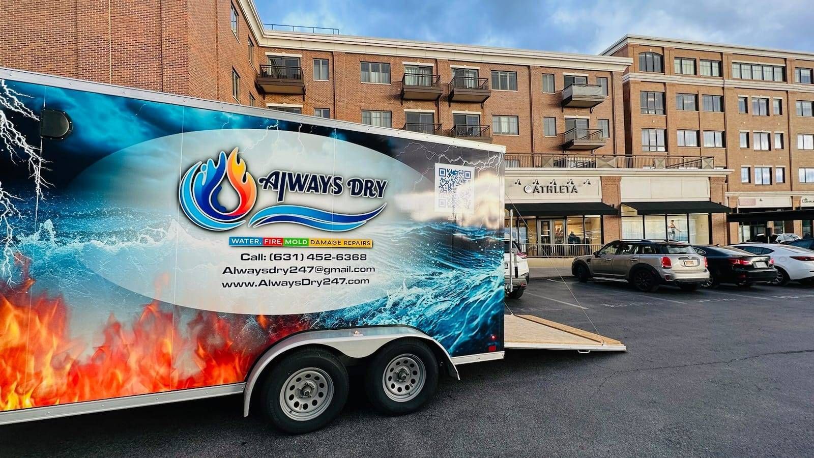 A branded Always Dry trailer with fire and water graphics parked in a parking lot in front of a brick commercial building.