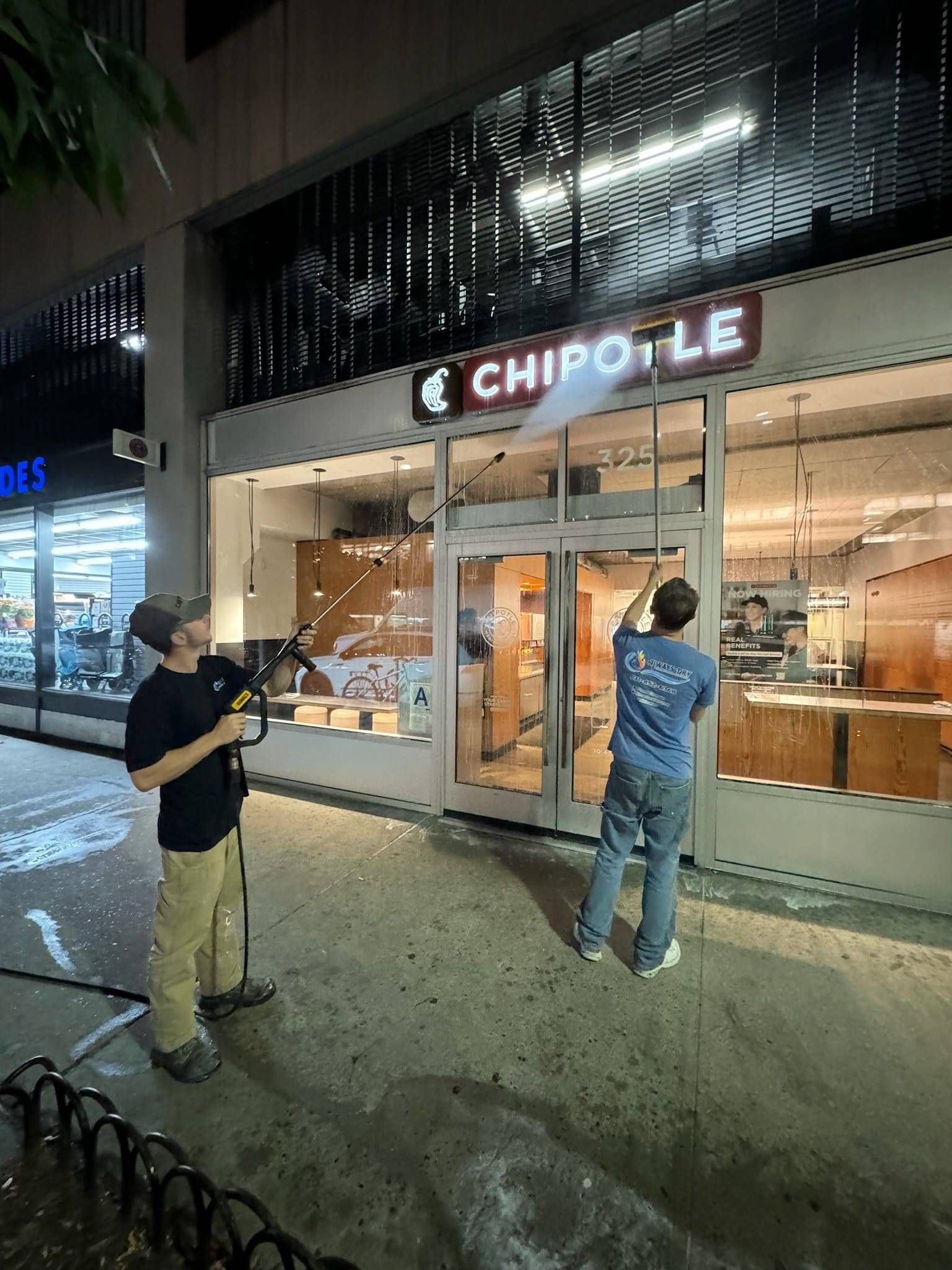 Two people use long-handled pressure washers to clean the front facade and glass doors of a Chipotle restaurant at night.