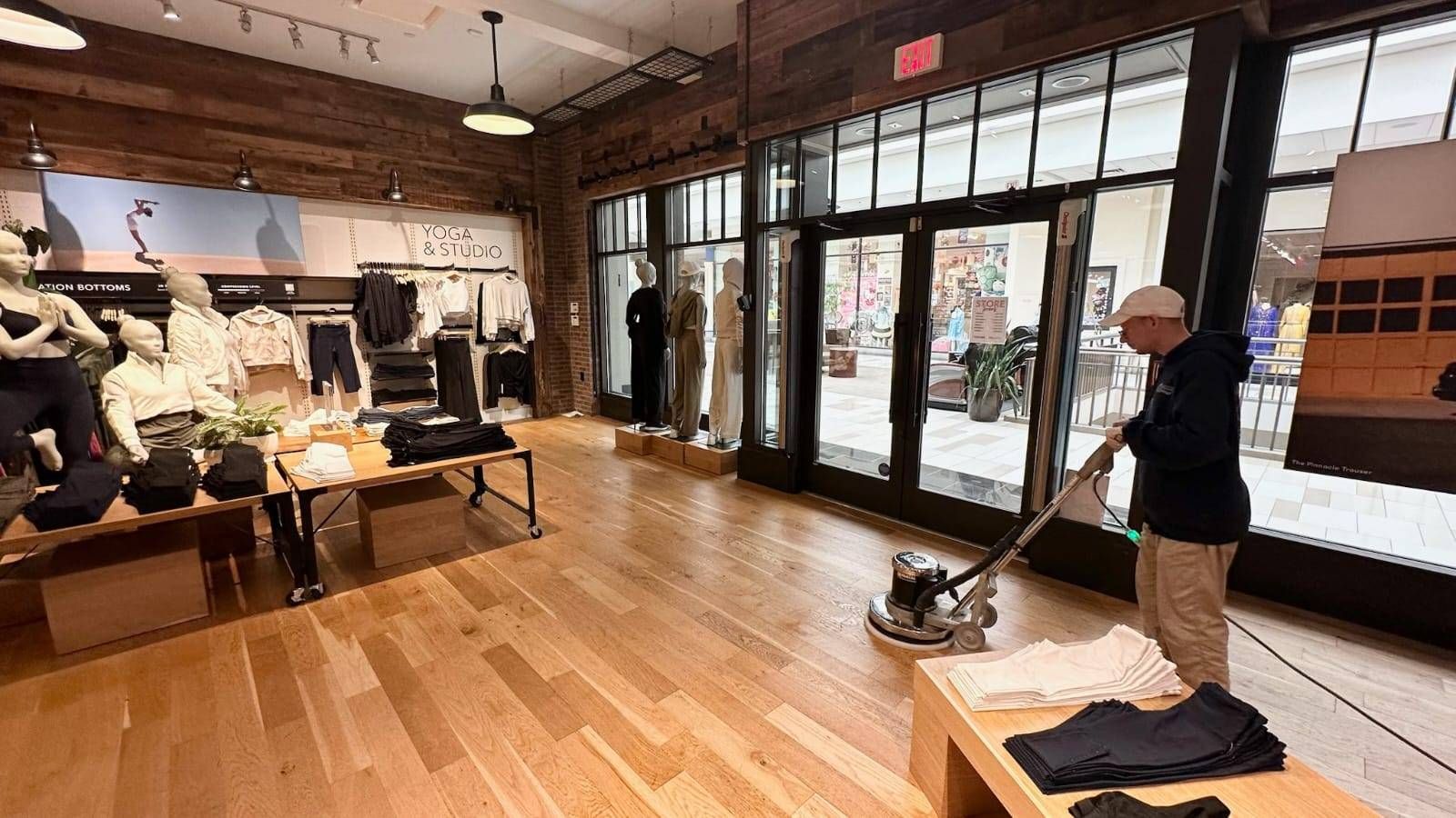 A person uses a floor buffer on the wooden floor of a retail clothing store featuring display tables and mannequins.
