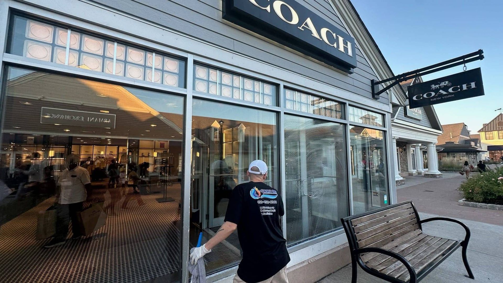 A person in a dark uniform cleans the large glass storefront of a Coach retail shop.