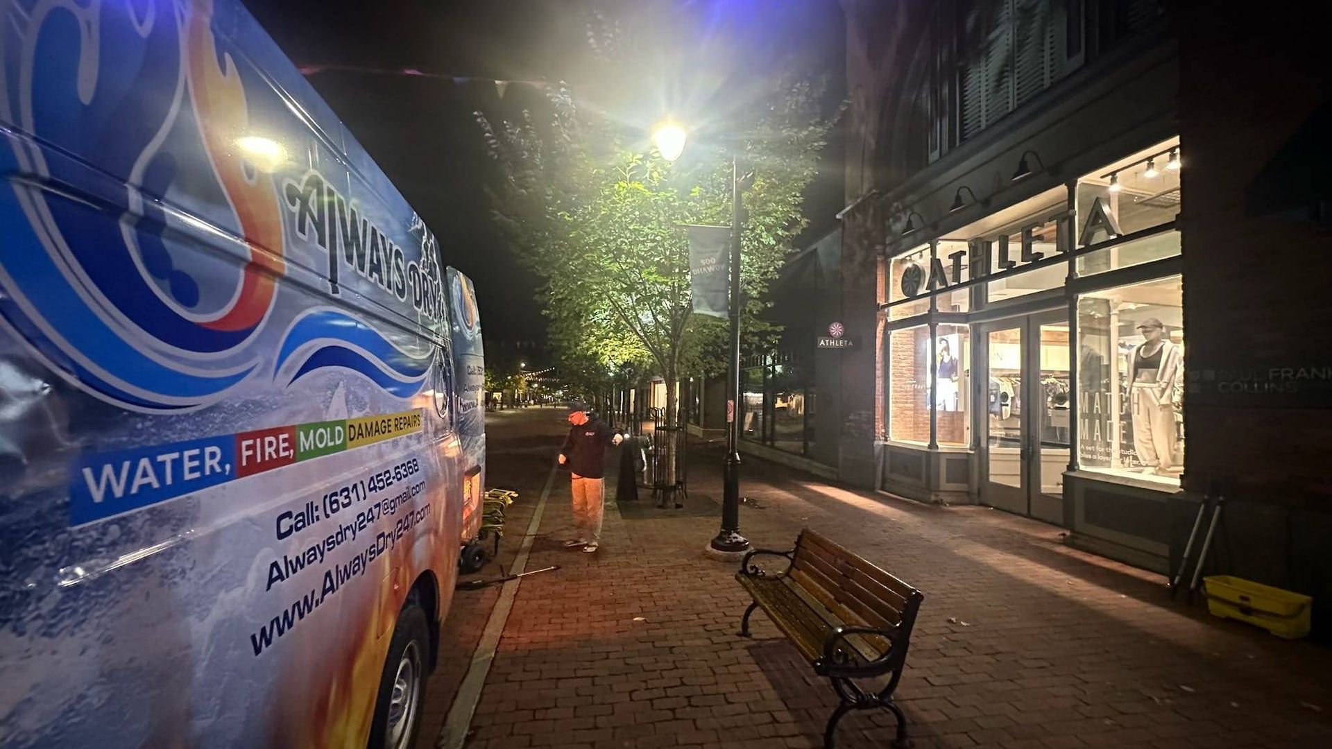 A brightly branded repair van parked on a brick sidewalk at night next to a lit storefront, with a bench nearby.