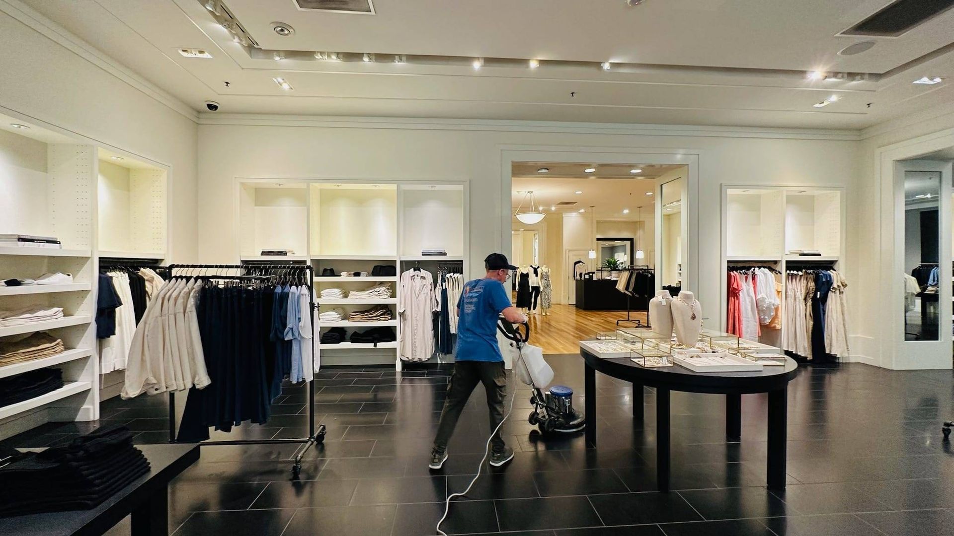A worker in a blue shirt uses a floor buffer to clean the dark tile floor of a brightly lit retail clothing store.