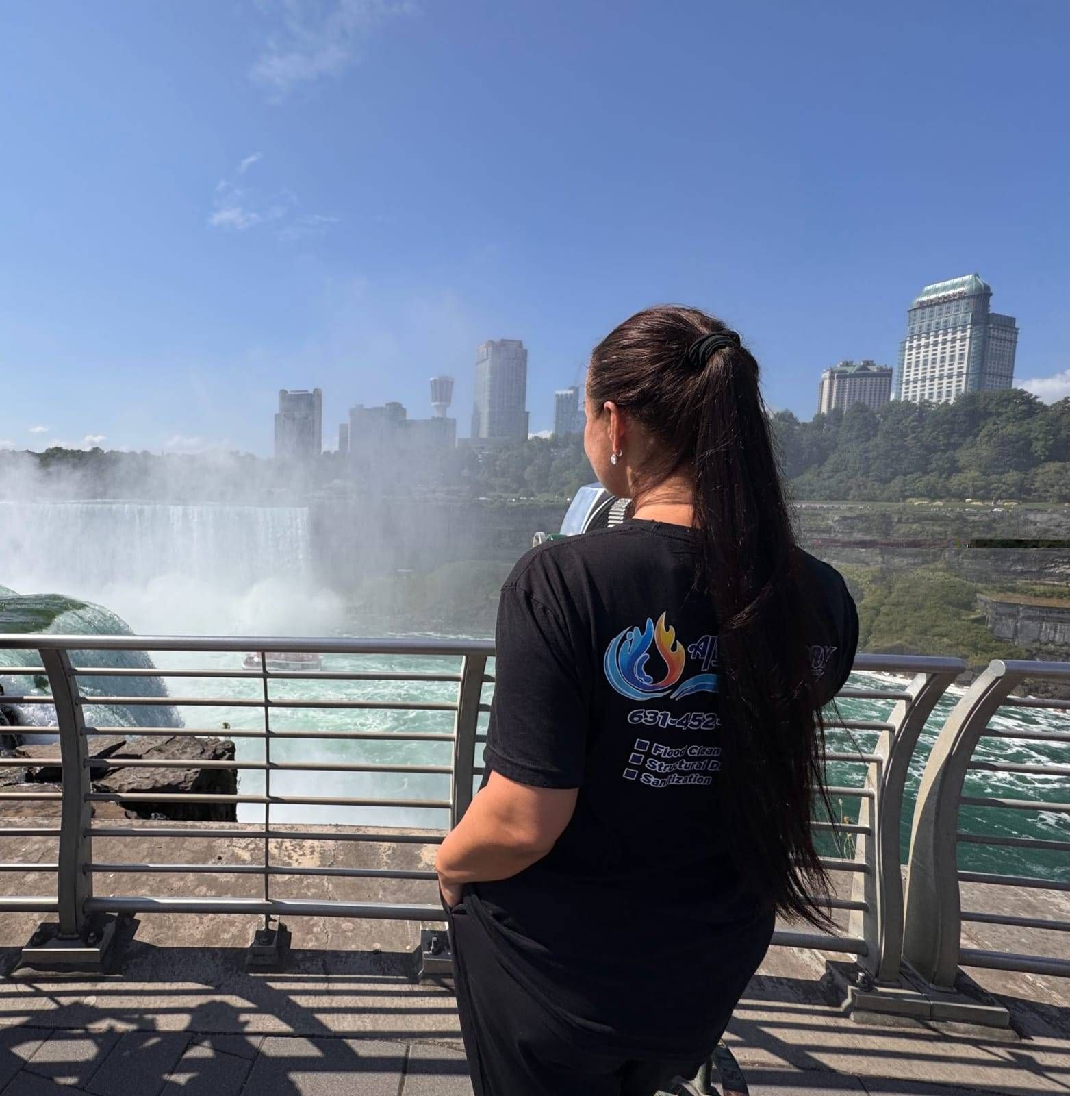 A person with long dark hair stands at a railing, looking out at the mist and buildings of Niagara Falls on a sunny day.